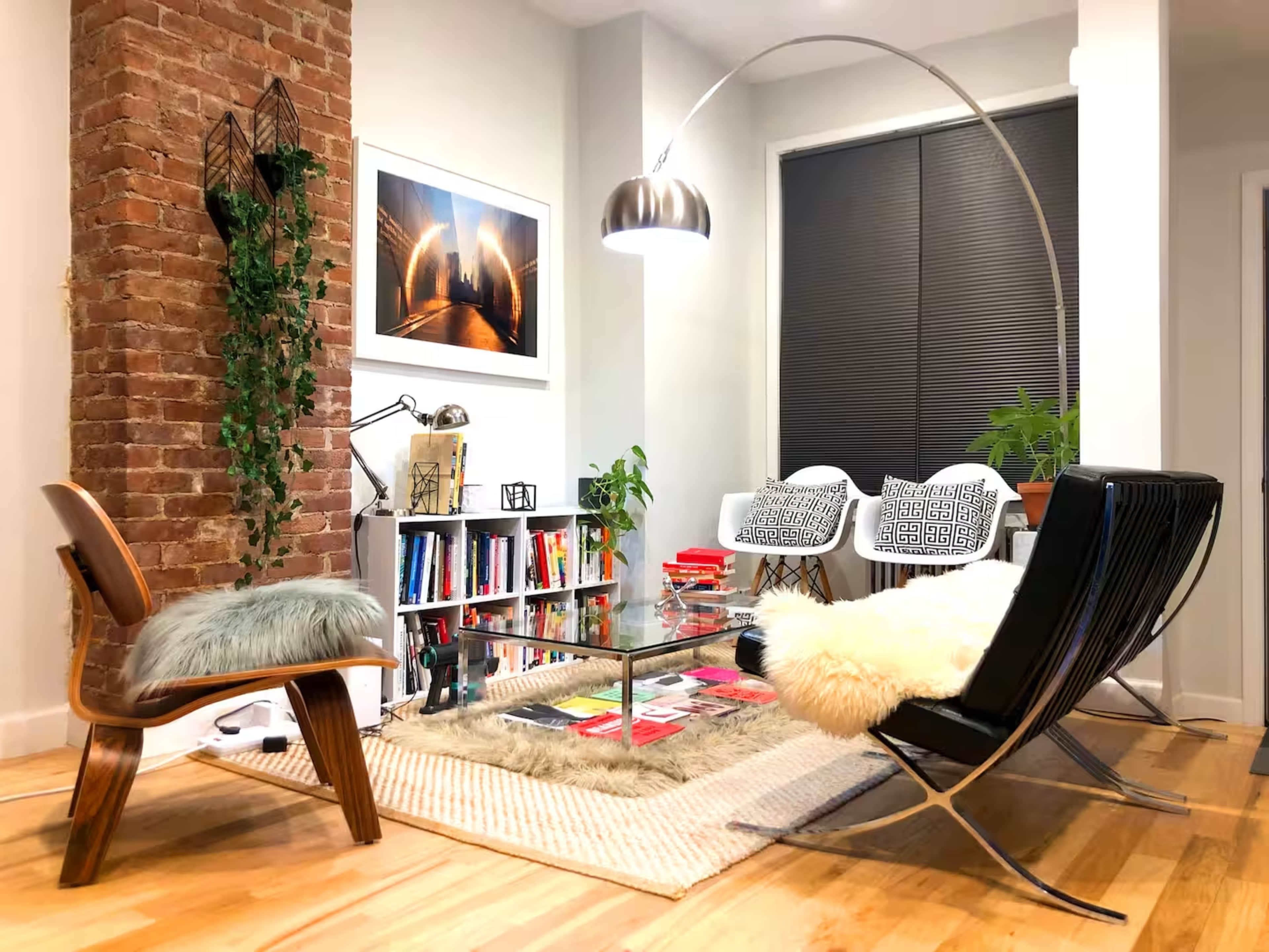 The image shows a modern living room with a brick accent wall, a glass coffee table, various books on a shelf, and two stylish chairs beneath an arc floor lamp.