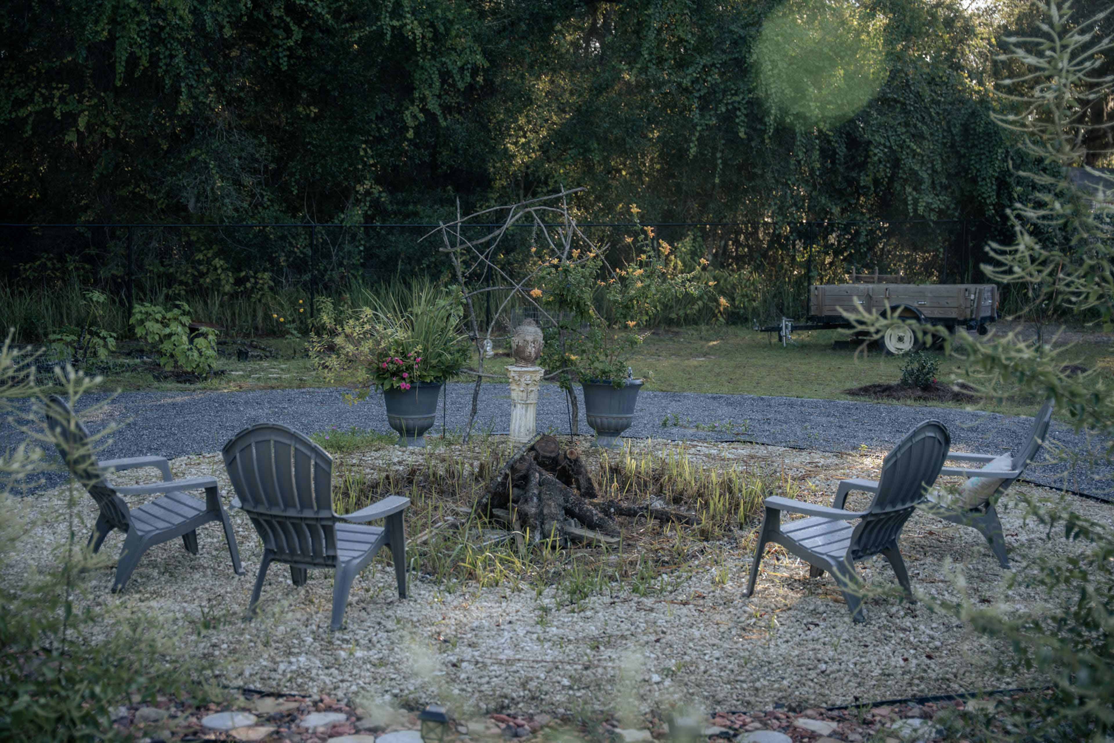 The scene shows a circular seating arrangement with four chairs facing a central fire pit, surrounded by gravel and potted plants in a garden setting.