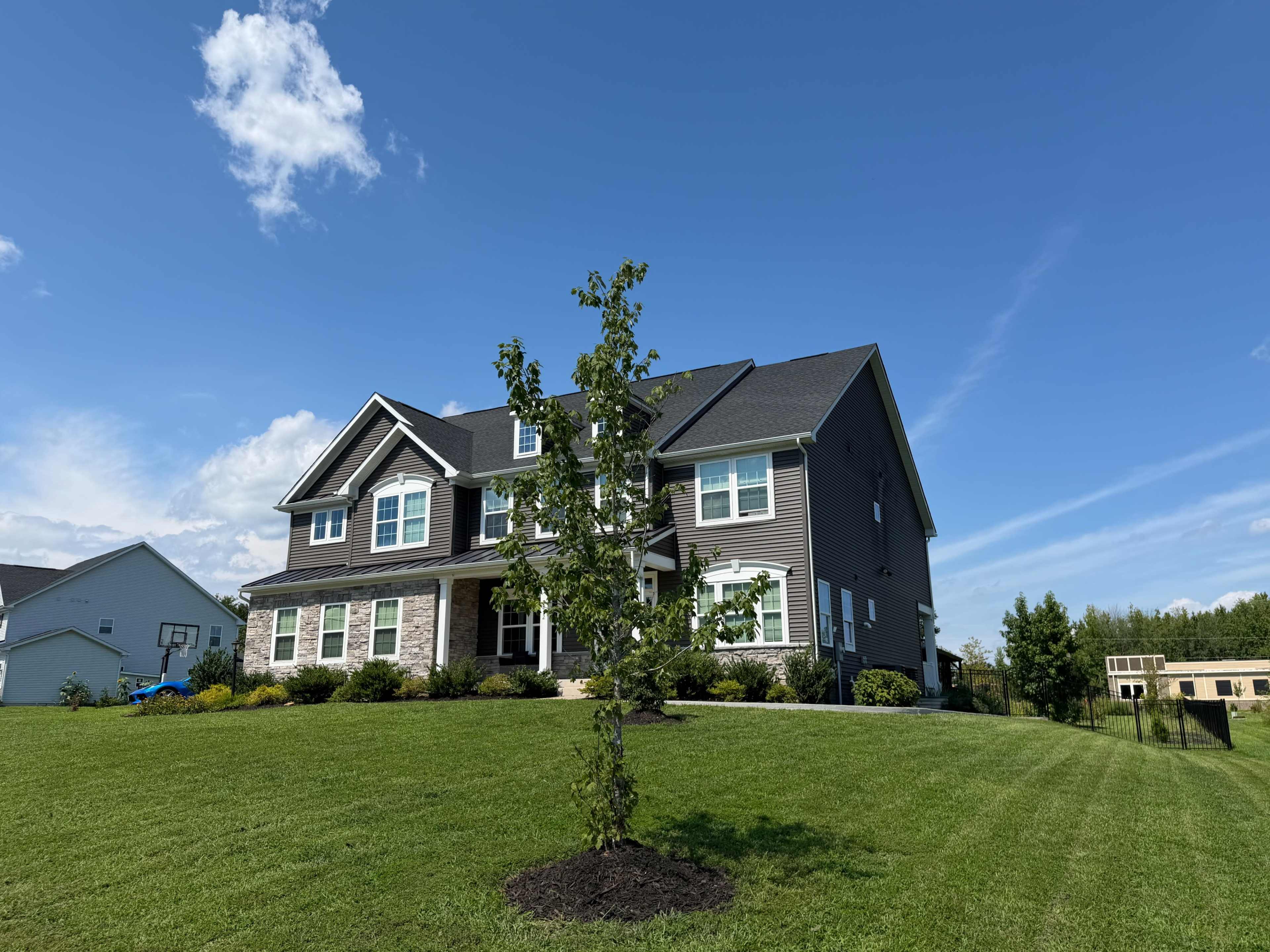 A large, two-story house with a dark exterior and green lawn is situated on a sunny day with a blue sky in the background.