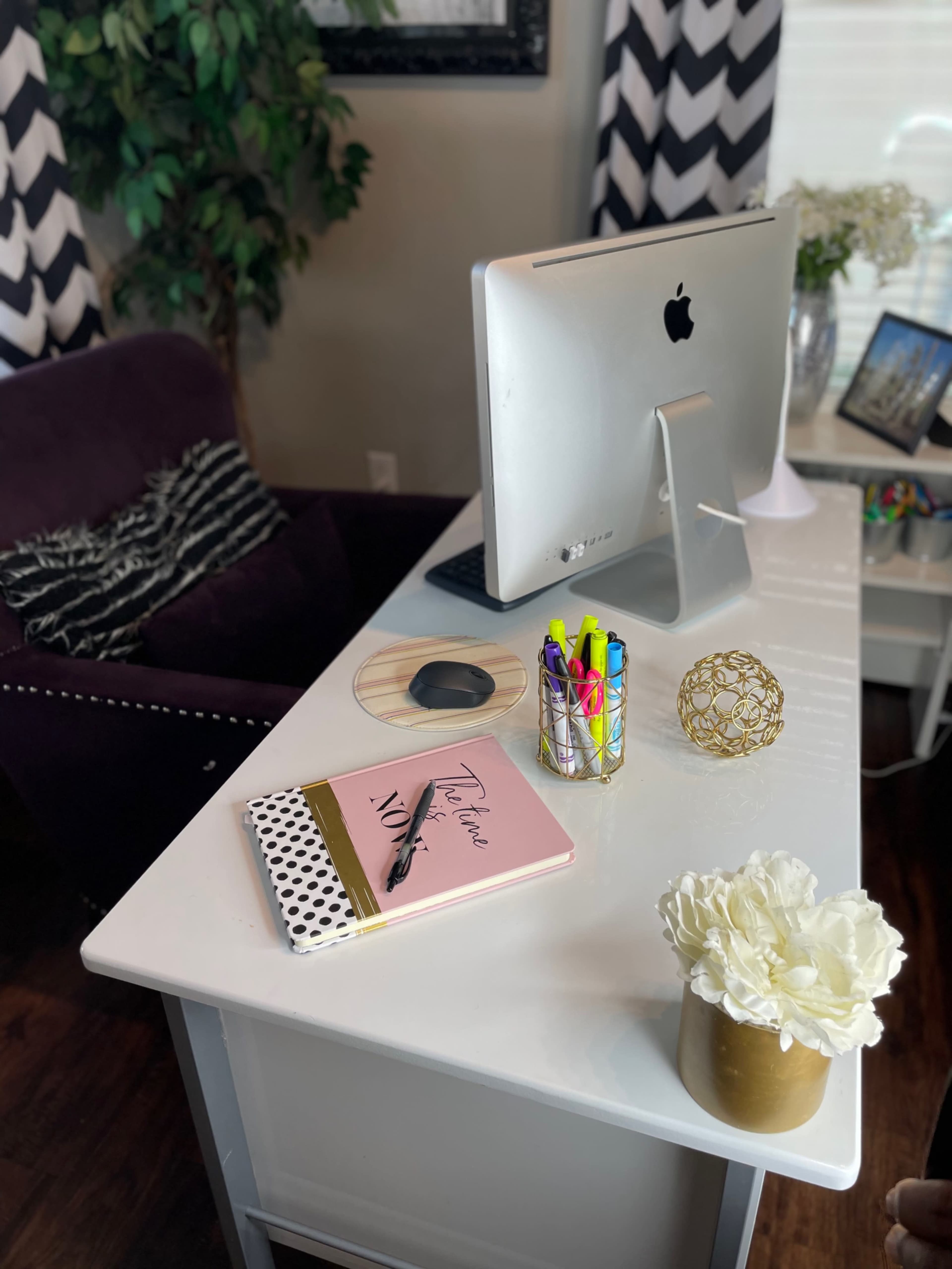 A modern desk setup features an iMac, colorful stationery, a decorative gold orb, a notebook, and a faux flower arrangement.