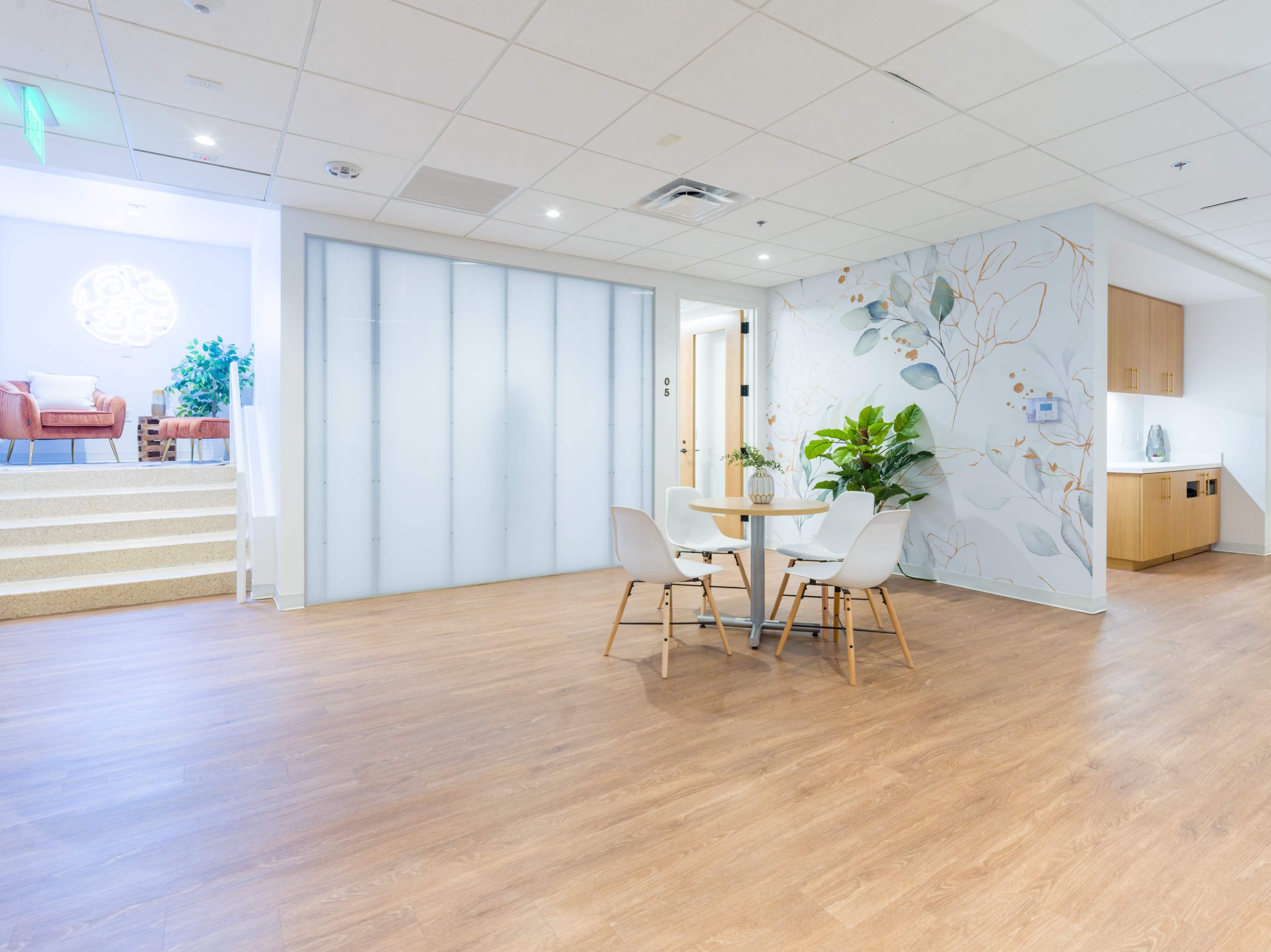 The image shows a modern, airy office space with a small seating area, a round table with four chairs, and a decorative wall featuring plants and abstract designs.