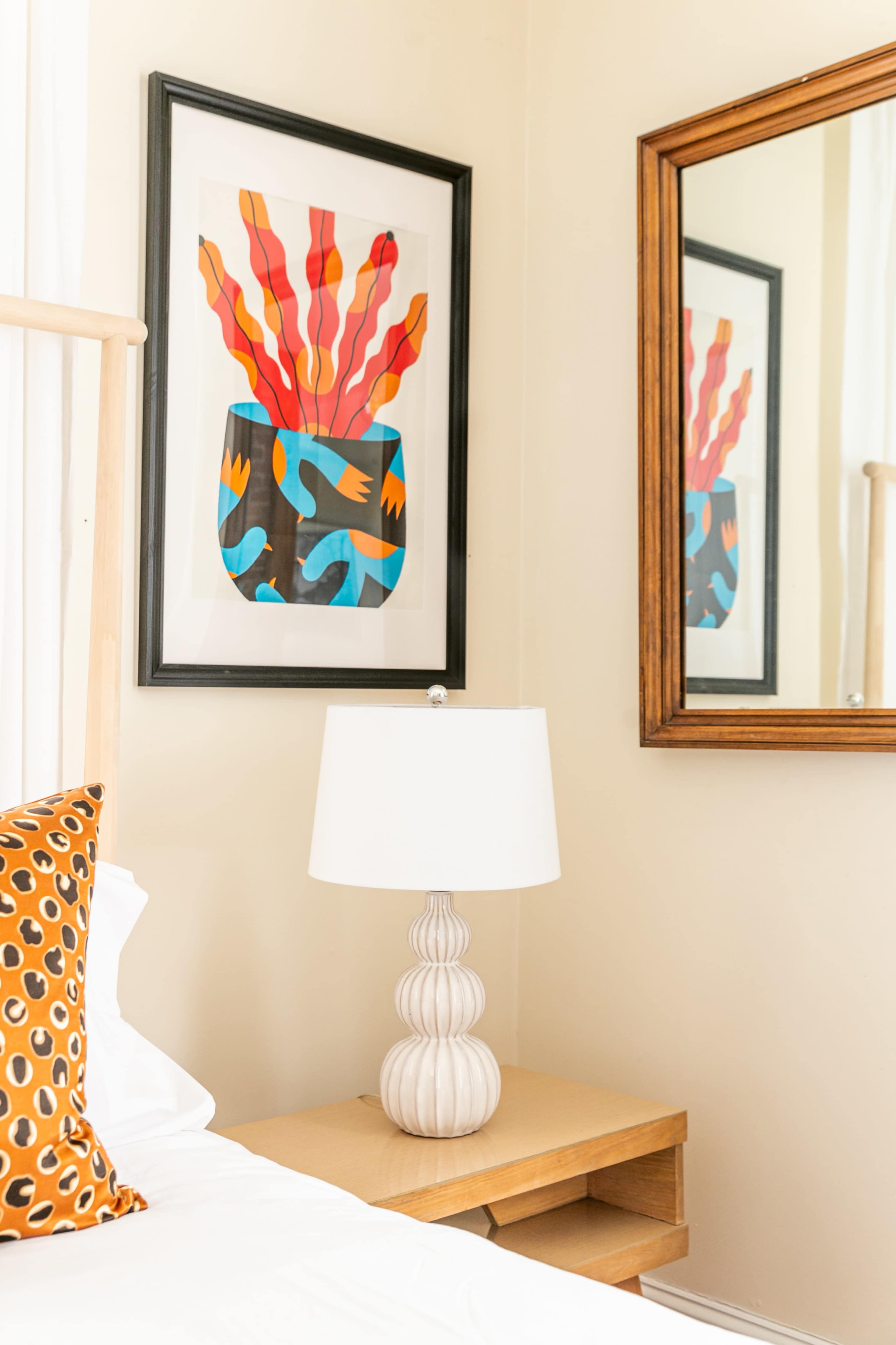 A bedside table with a lamp and a framed piece of abstract art mounted on the wall next to a mirror.