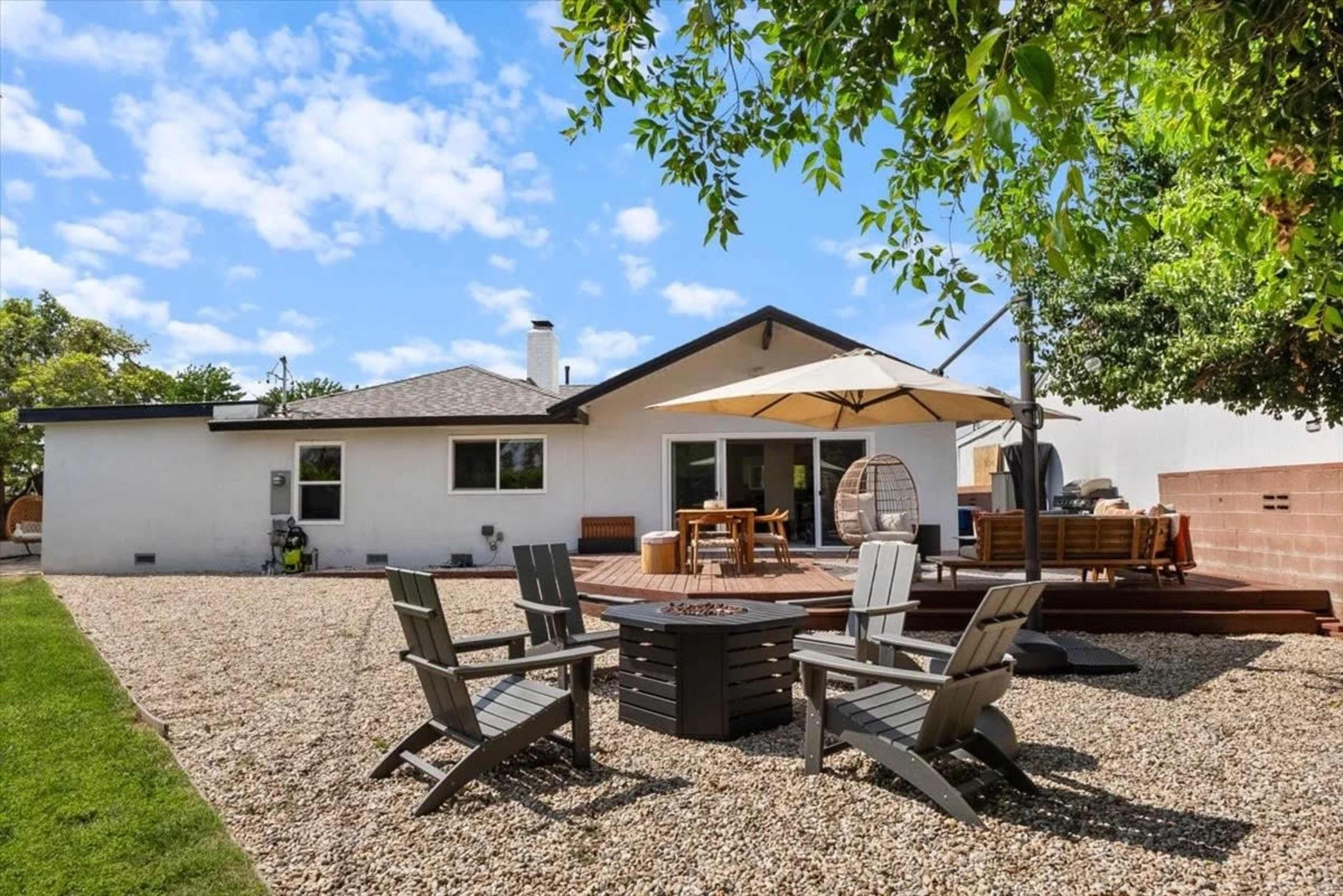 The image shows a backyard with gravel, seating areas, a wooden deck, and a house with a large umbrella overhead.