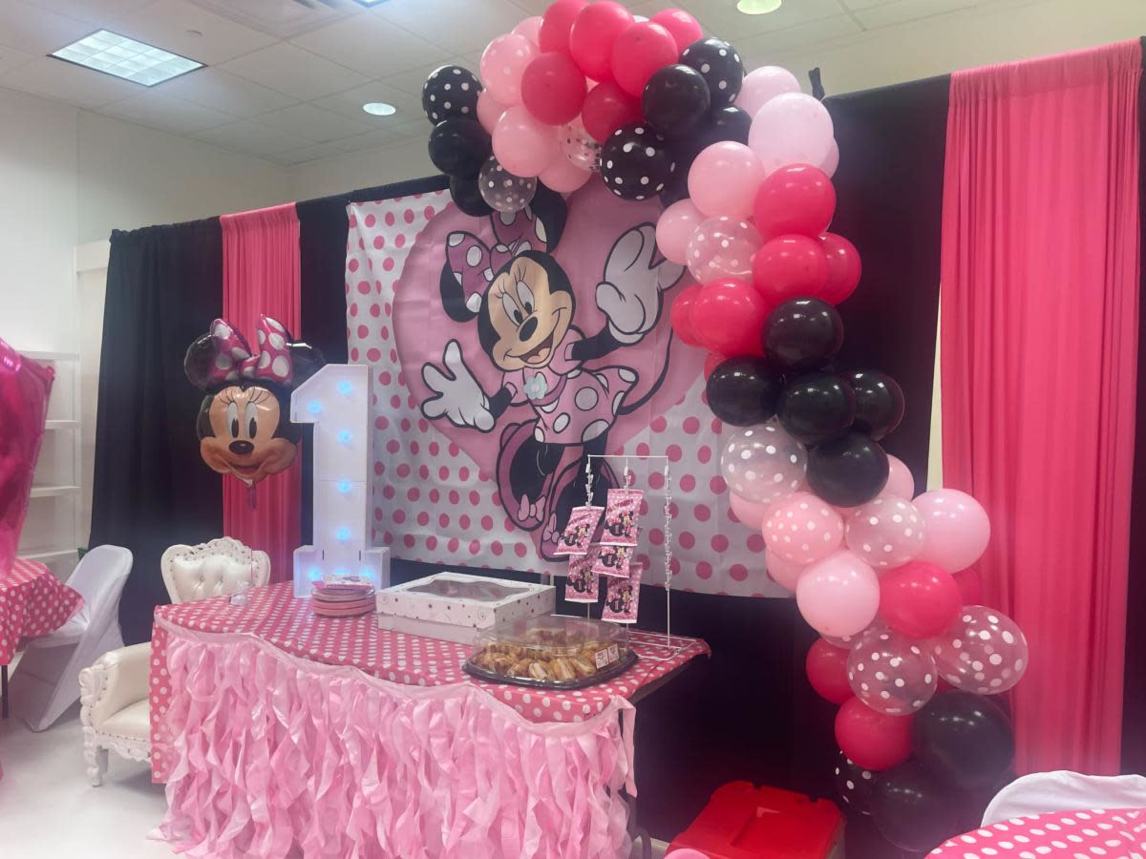 The image shows a birthday party setup featuring a large Minnie Mouse backdrop, a decorative balloon arch in pink and black, and a table with a cake display and treats.