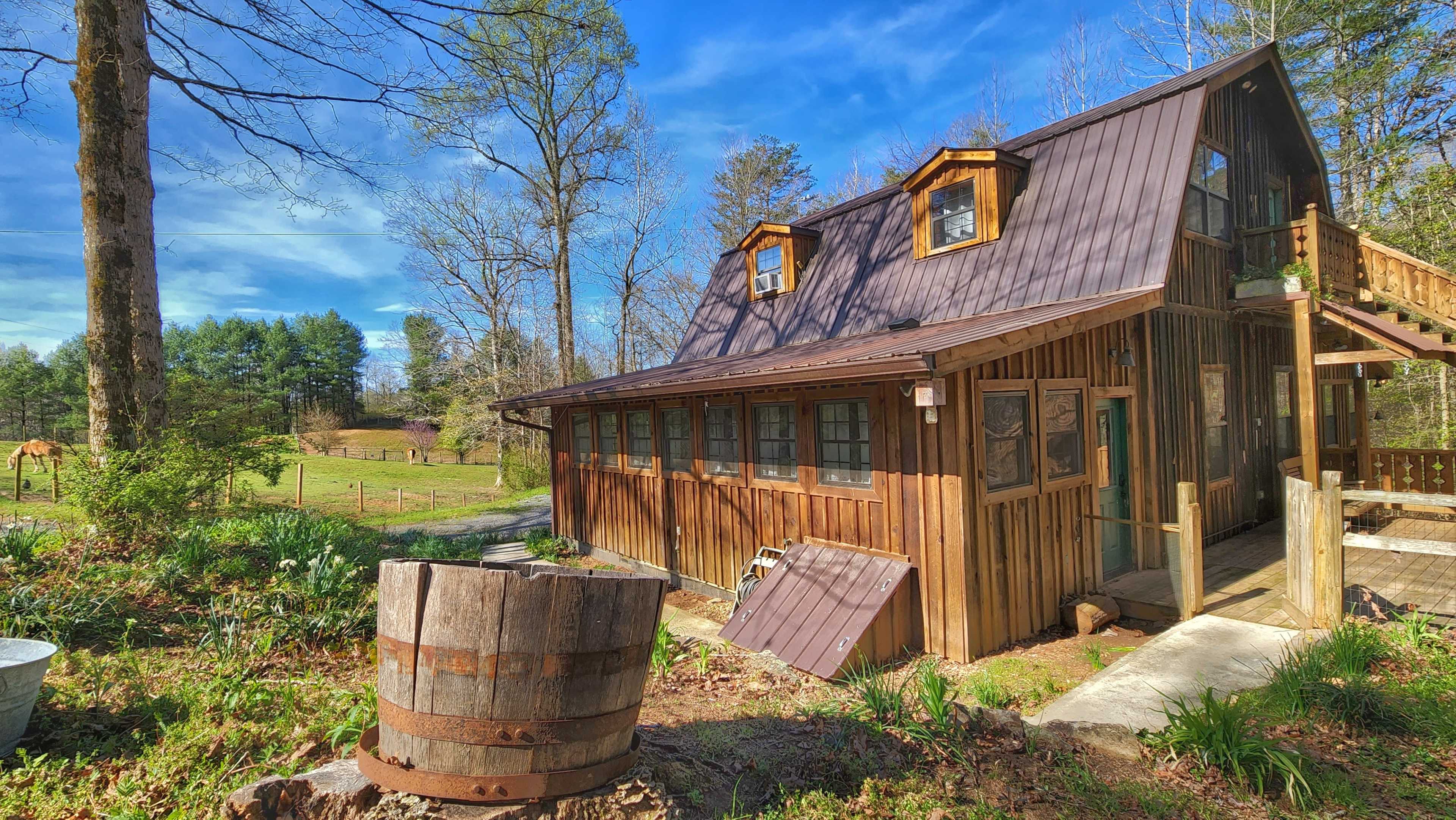 A wooden cabin with a steep roof and large windows sits beside a wooden barrel in a wooded area.