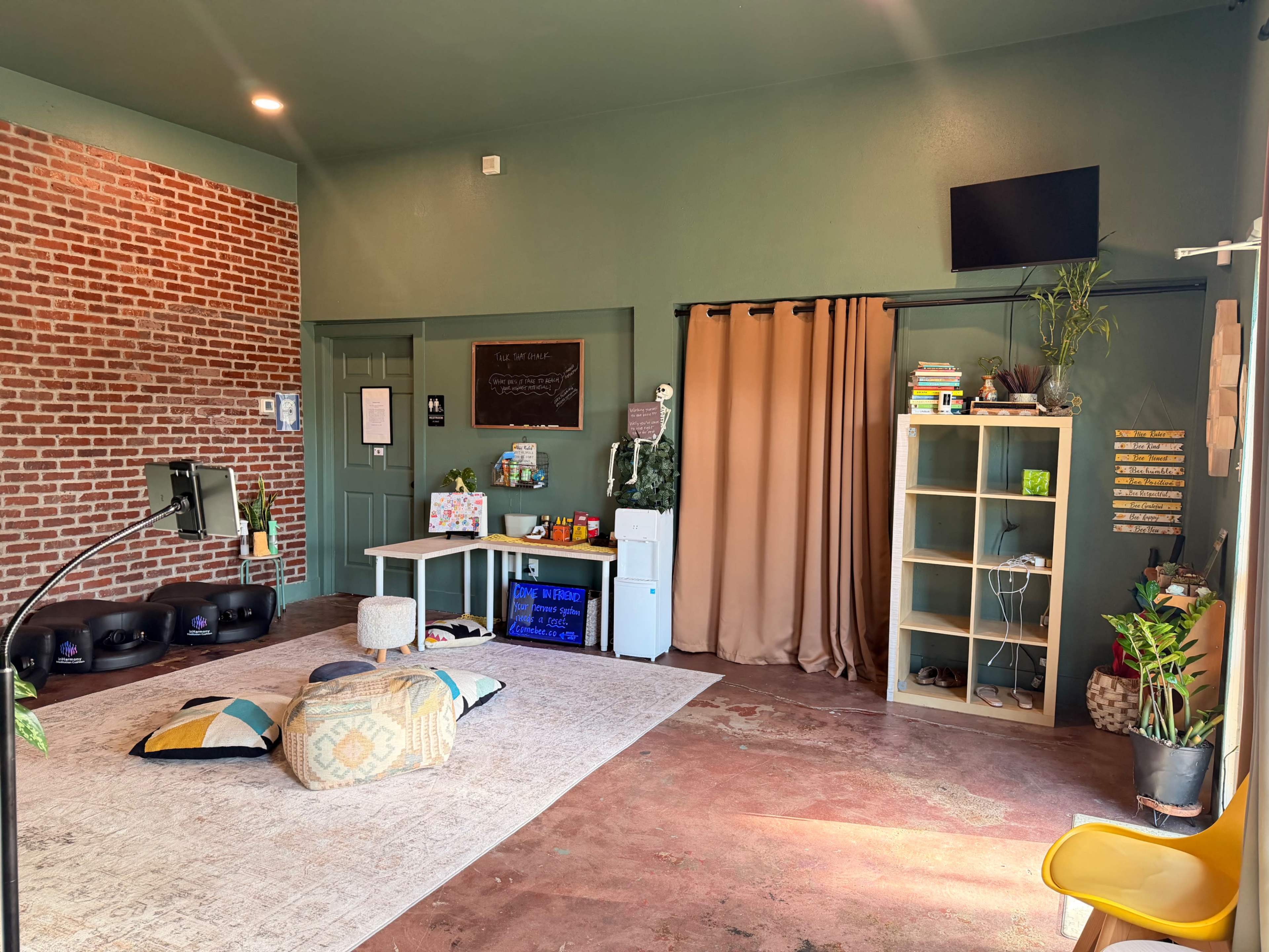 The image shows a spacious room with brick walls, a large area rug, a seating area with cushions, a desk with a chalkboard, and a shelf filled with books and plants.