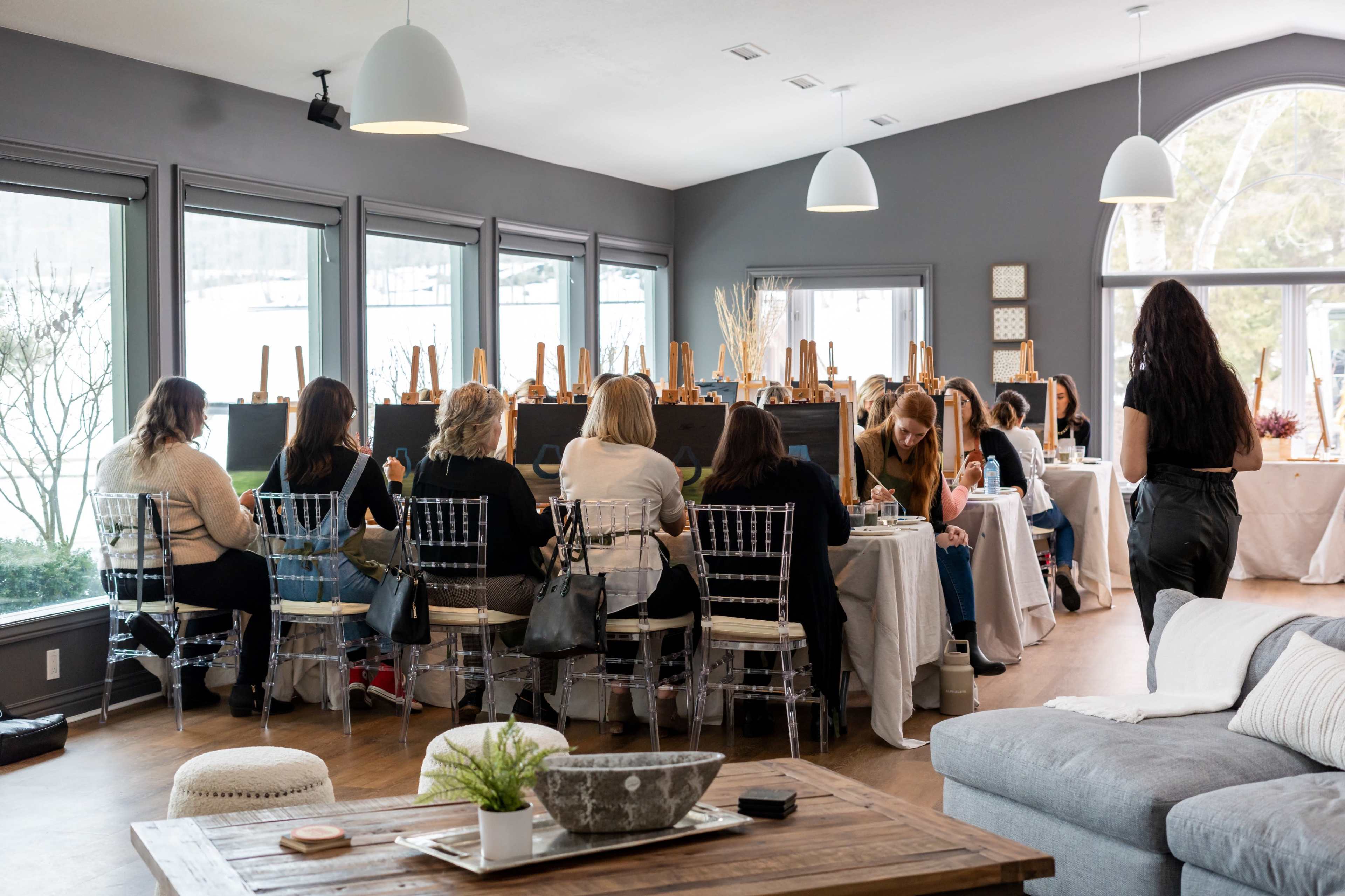 A group of people is seated at long tables in a bright and airy art studio, each with an easel in front of them.