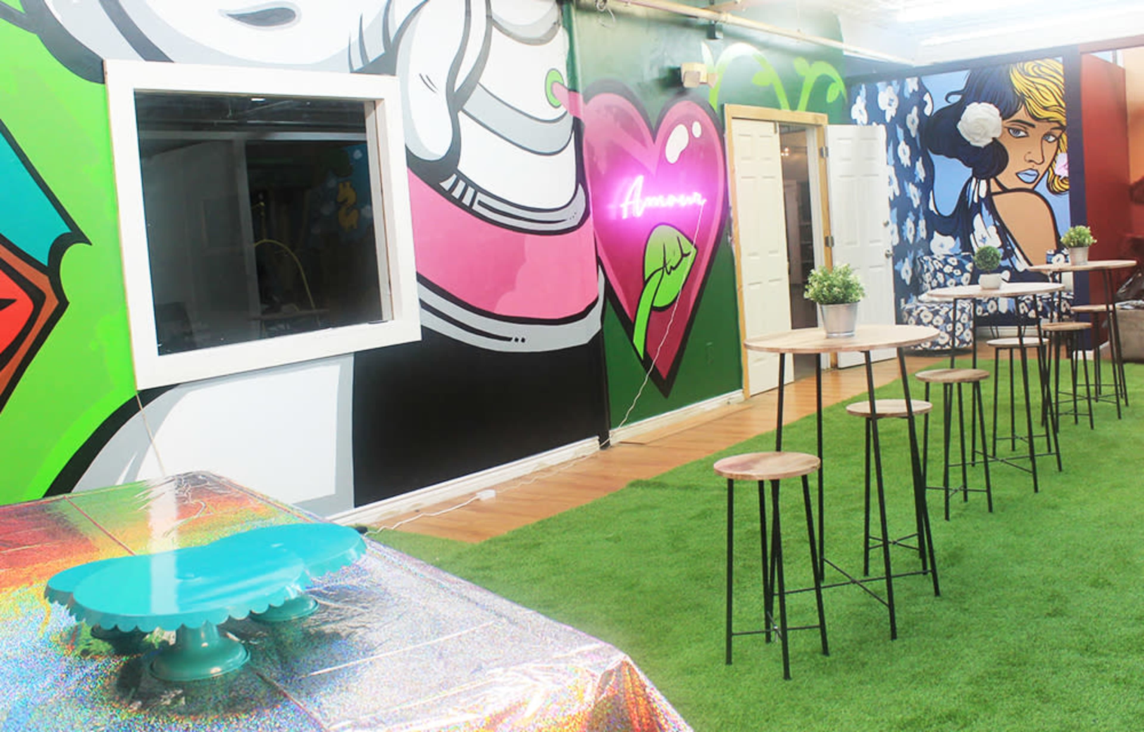 The scene features a vibrant indoor space with colorful graffiti on the walls, a green grass-like floor, and several high wooden tables surrounded by stools.