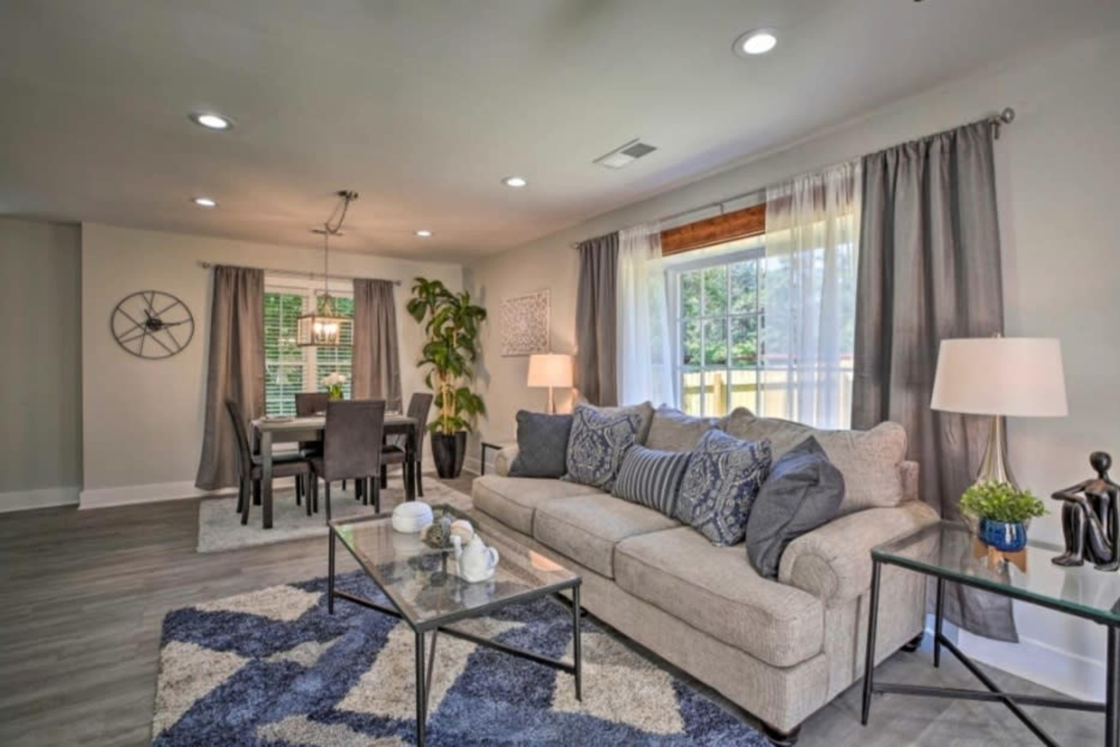 The living room features a light-colored sofa with decorative pillows, a glass coffee table, and a dining area with a chandelier visible in the background.