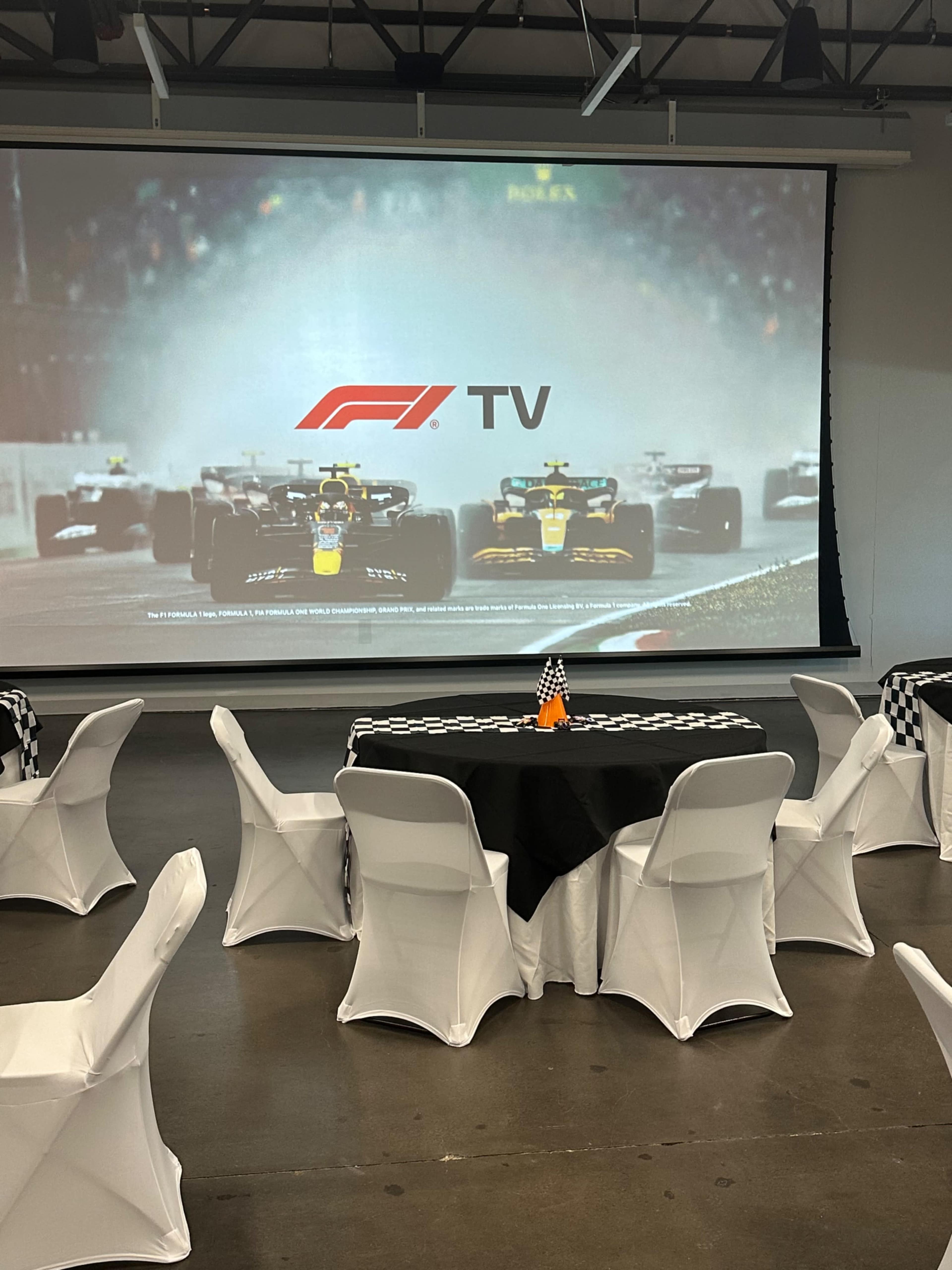 A racing-themed event space features round tables with checkered tablecloths and a large screen displaying an F1 TV broadcast, showcasing multiple race cars.