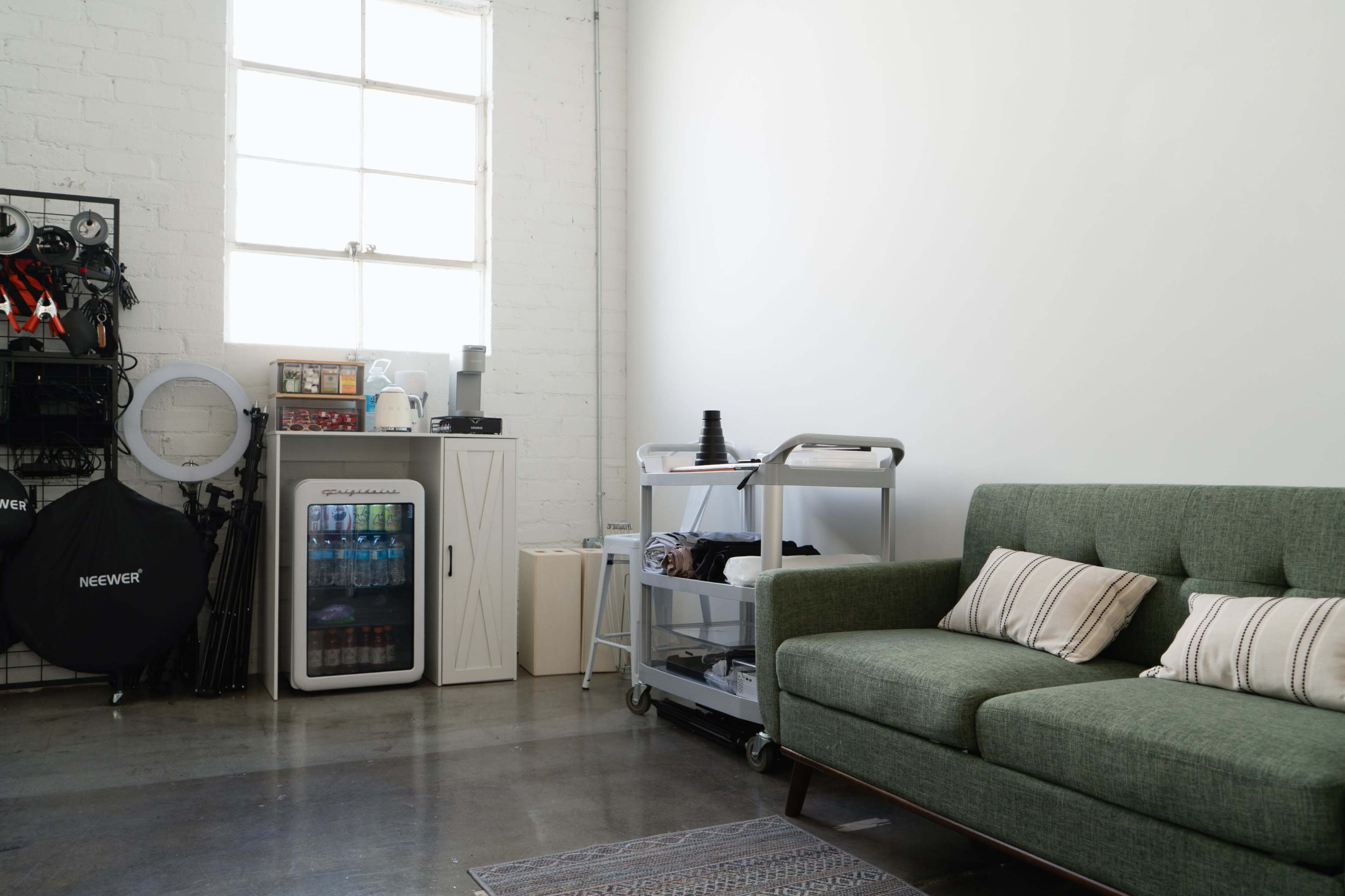 The image shows a minimalist room featuring a green sofa, a cart with various items, a small refrigerator, and a window allowing natural light to enter.