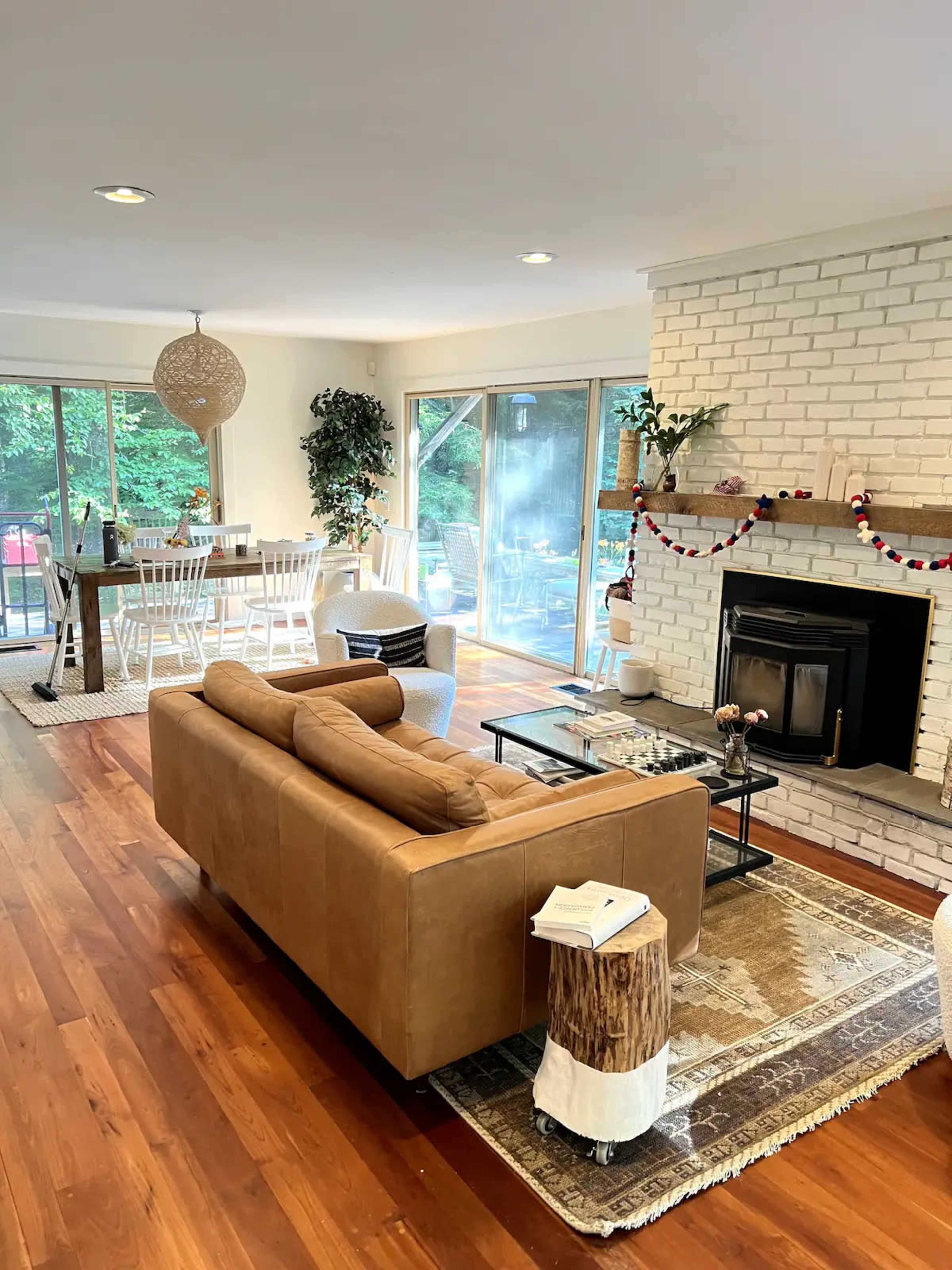 A living room with a brown couch, a coffee table, and a white brick fireplace, leading to a dining area with a wooden table and white chairs.