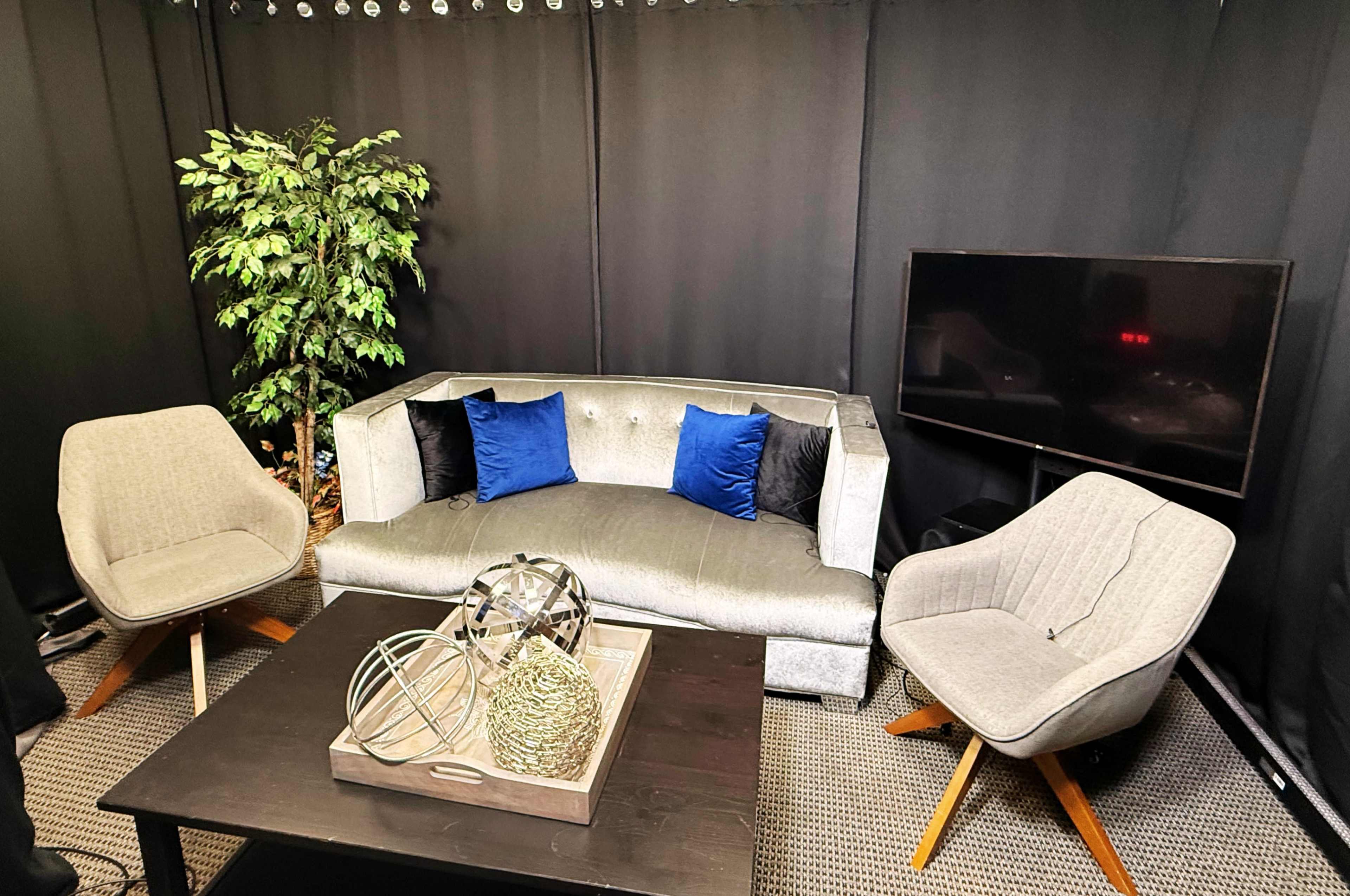 The image shows a small indoor seating area with a gray couch, two upholstered chairs, a coffee table with decorative items, and a television against a black backdrop.