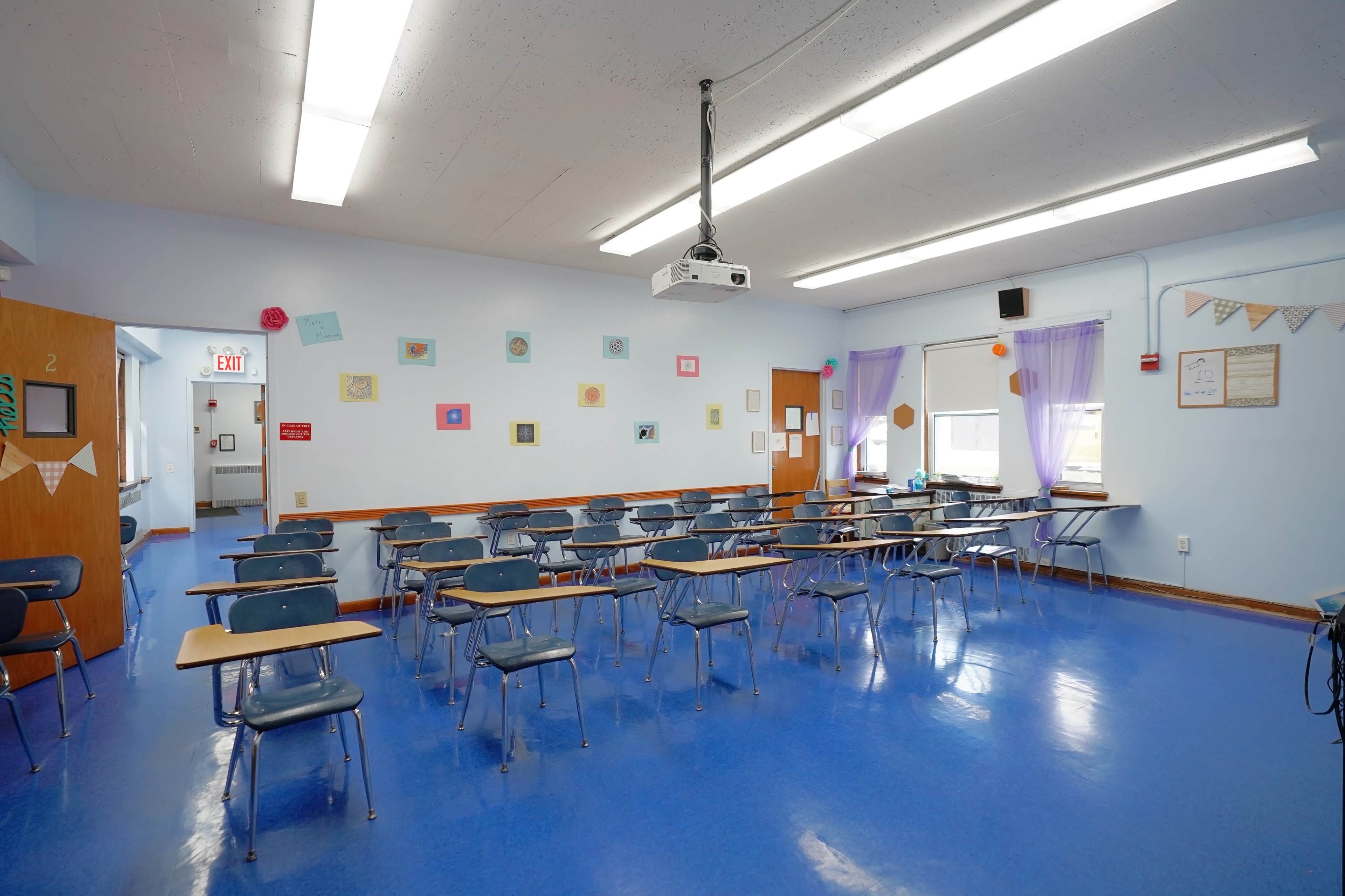 A classroom with blue floors and walls, containing rows of desks, a projector on the ceiling, and wall decorations.