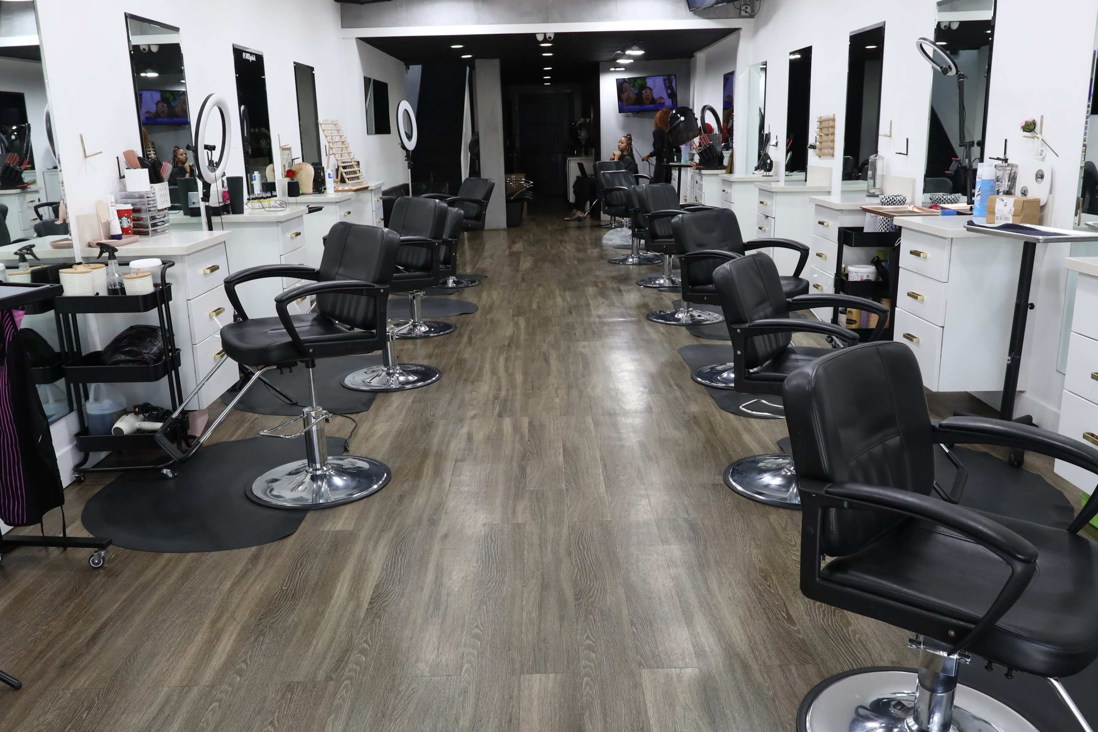 A spacious hair salon with several styling stations, each equipped with a black chair and large mirrors, located on either side of a wooden floor aisle.