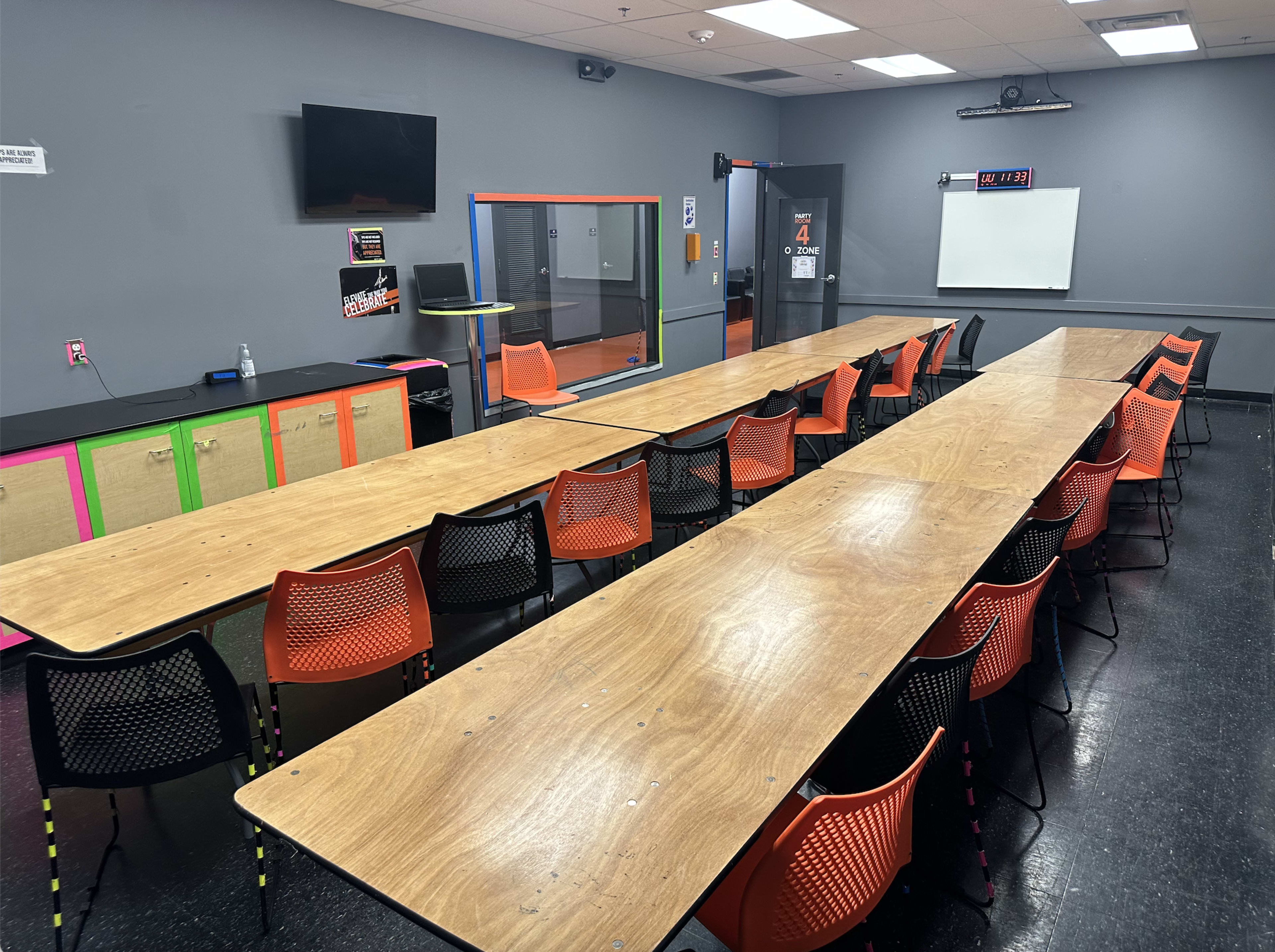 A spacious classroom with multiple wooden tables arranged in rows and colorful chairs, along with a television and whiteboard on the walls.