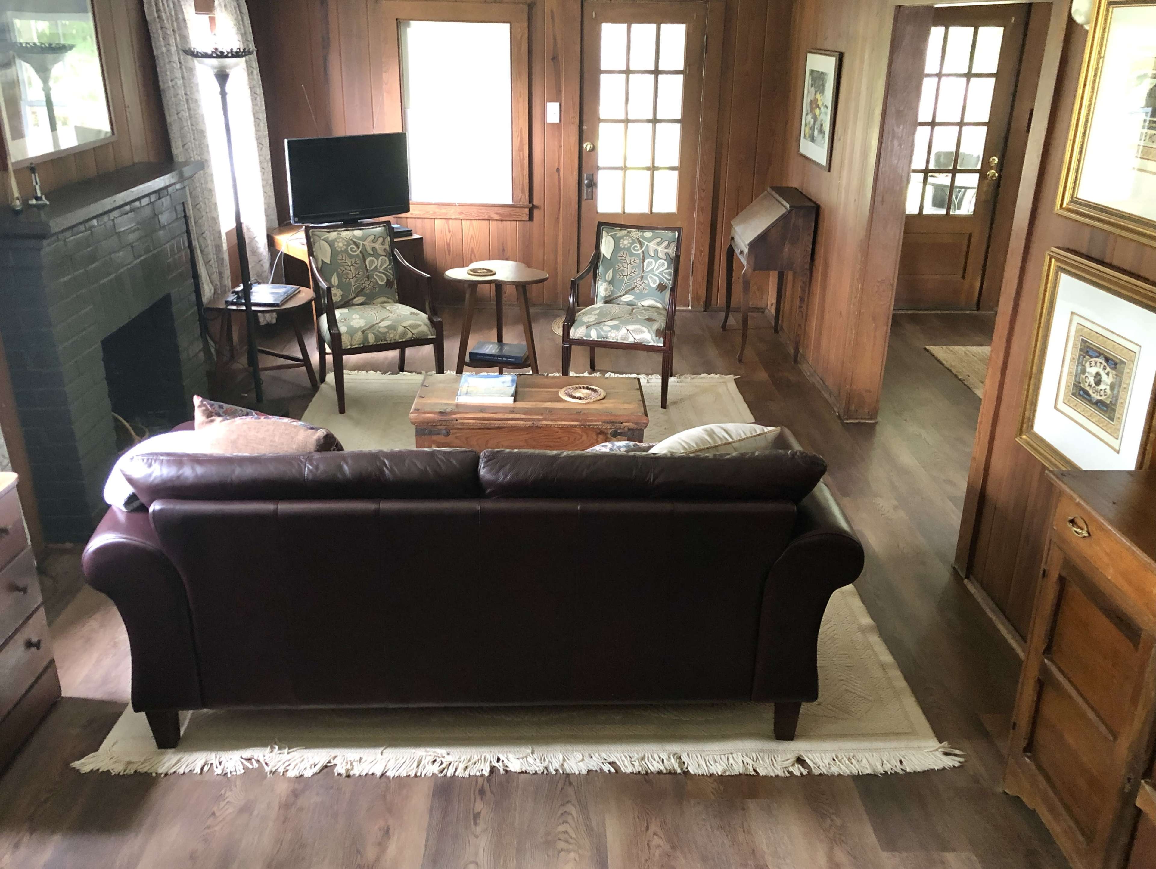 A living room features a dark leather sofa facing two patterned chairs and a wooden coffee table, with a television on a stand against a wall.