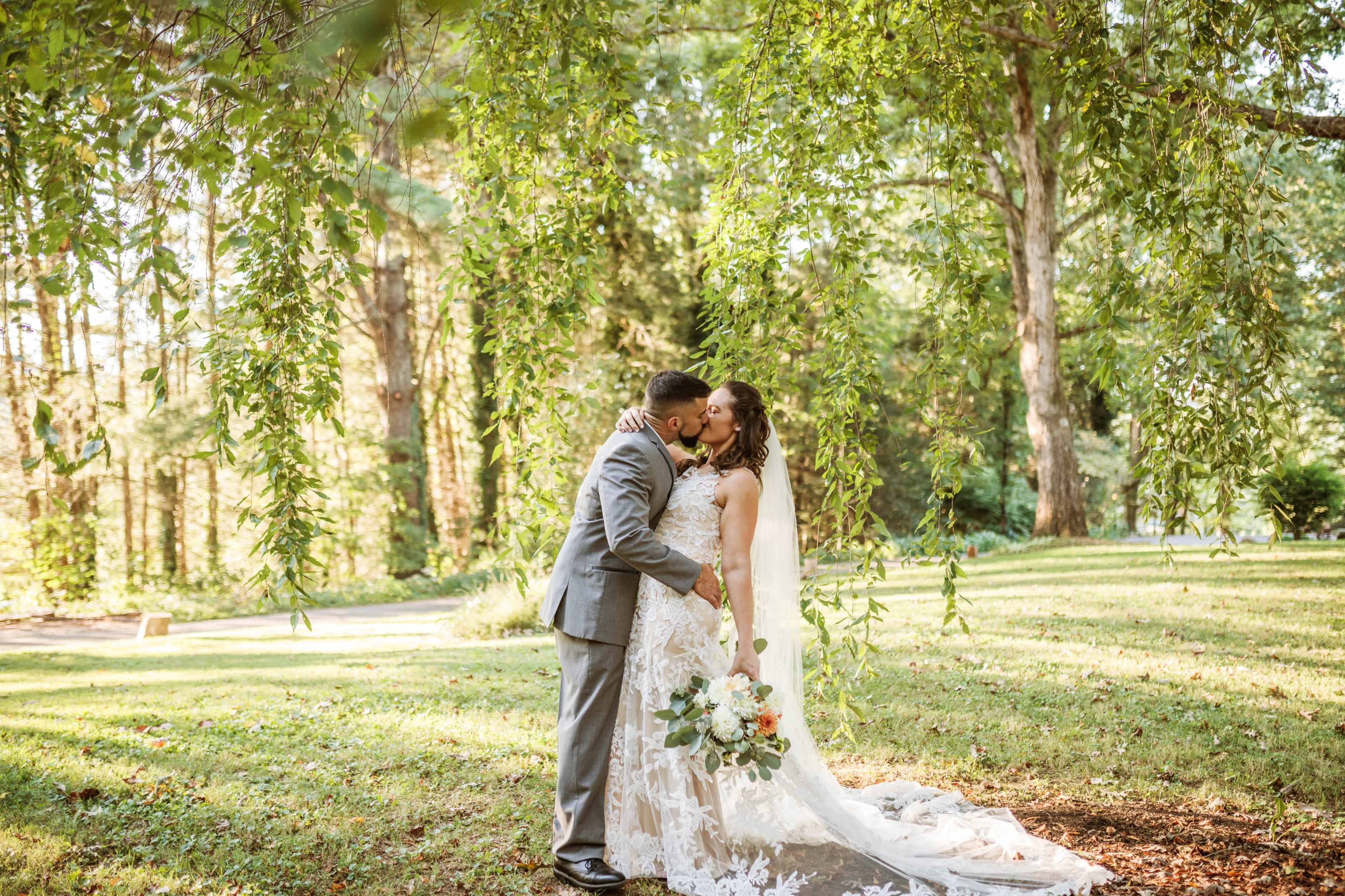 A couple exchanges a kiss under leafy trees in a garden setting during their wedding.