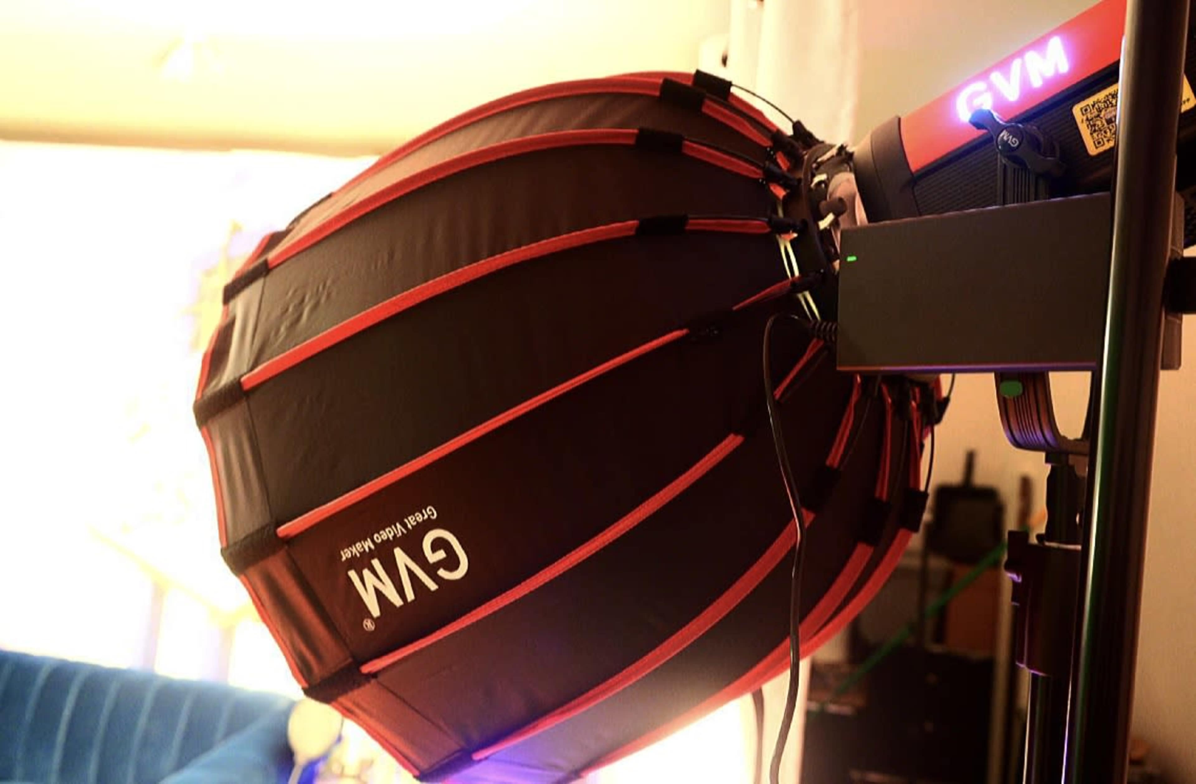 A large softbox with a red and black design is mounted on a lighting stand, positioned to illuminate a scene.