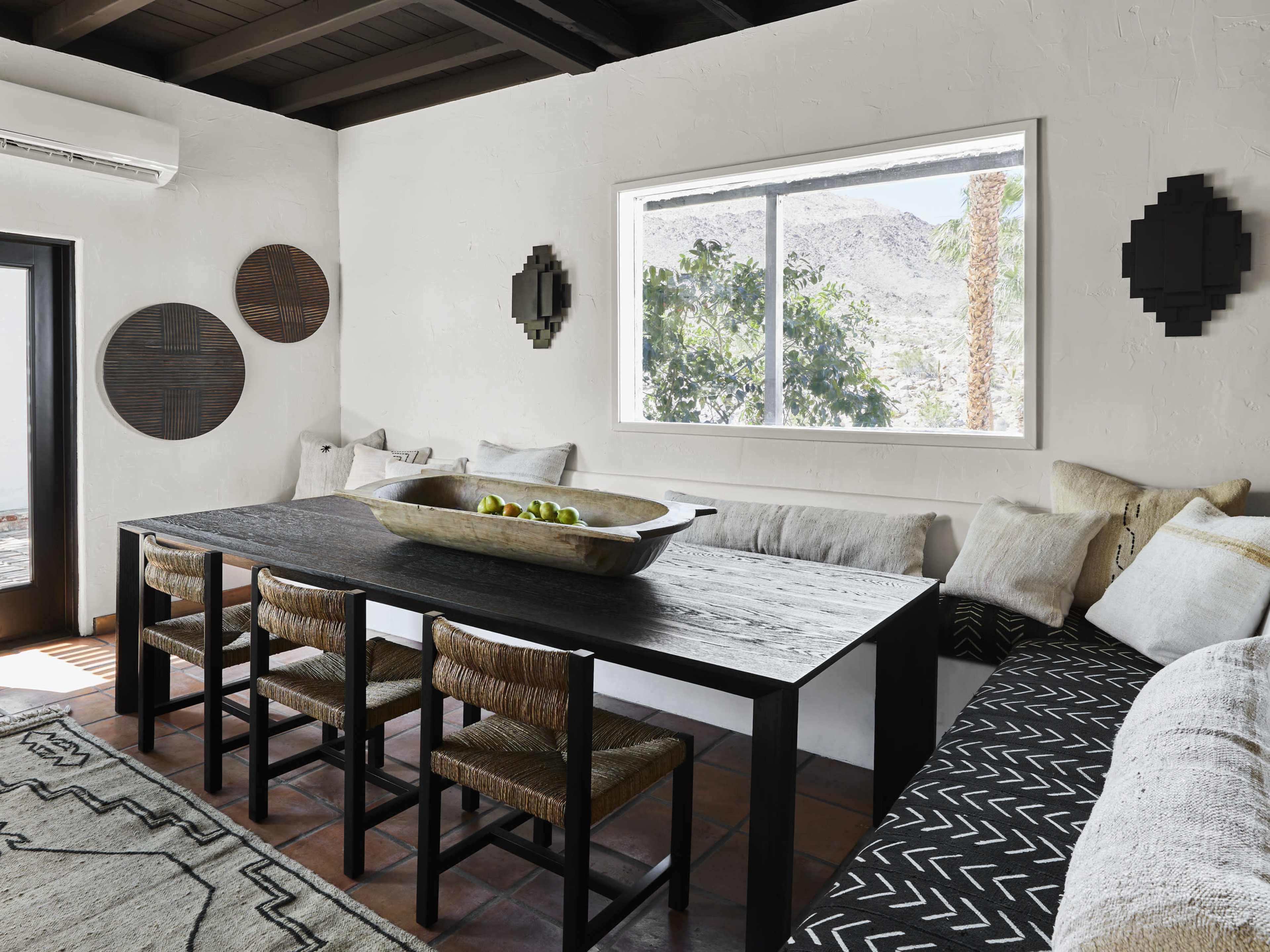 The image shows a modern, minimalist dining area with a large black table, woven chairs, and a cushioned bench against a white wall, featuring a window that overlooks a desert landscape.