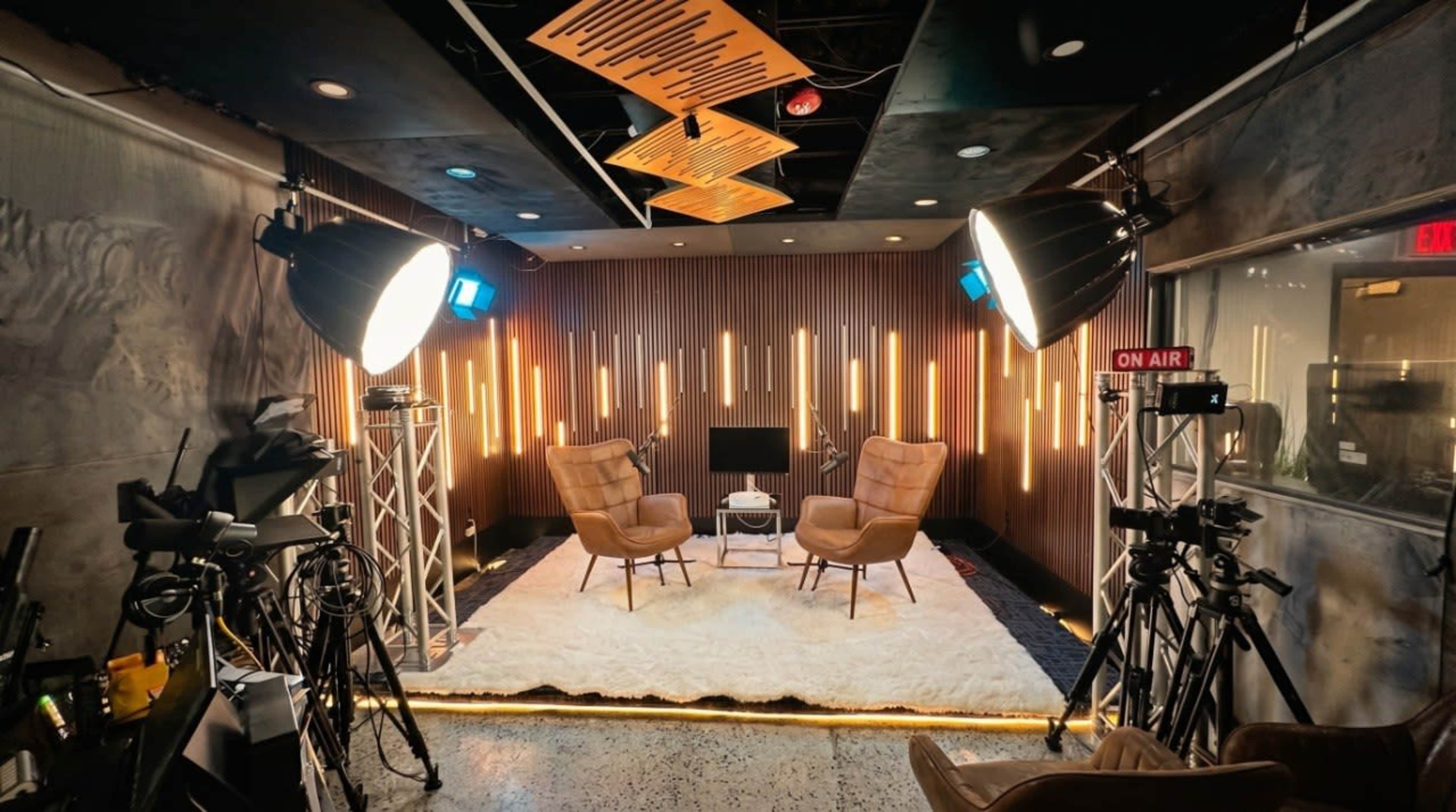 The image shows a modern studio setup with two armchairs facing a central table, surrounded by professional lighting and camera equipment.
