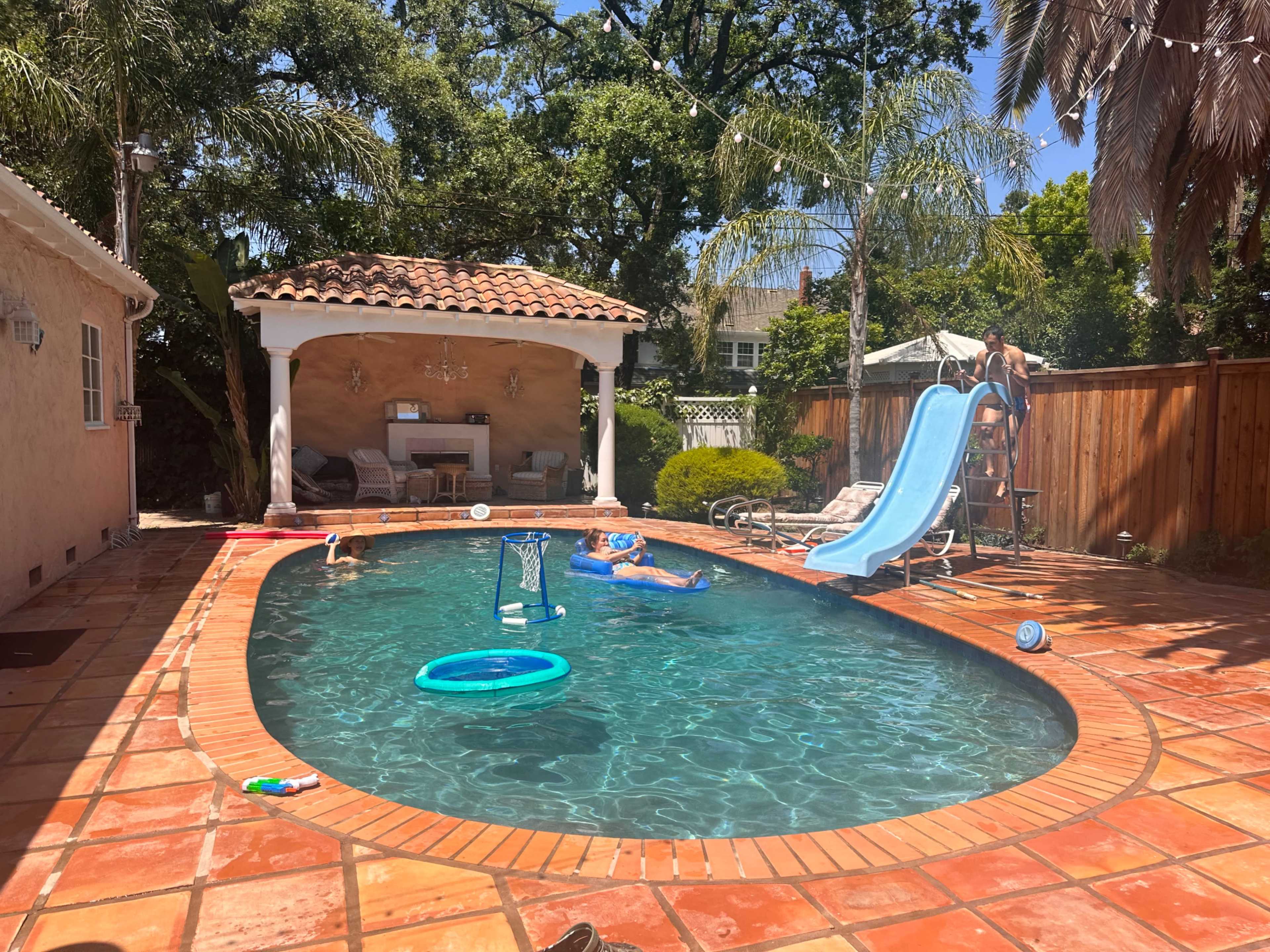 A backyard swimming pool features a slide, floating toys, and lounging areas surrounded by lush greenery and a covered seating area.