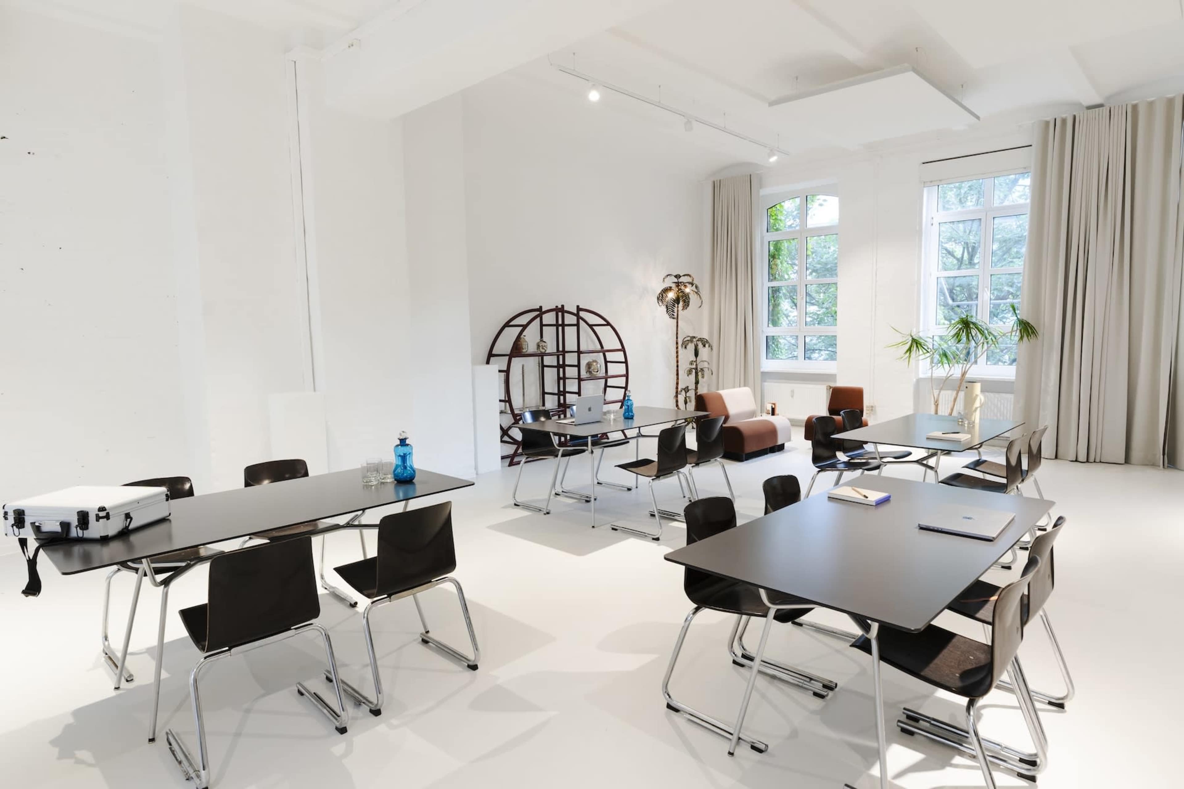 The image shows a well-lit, modern room with multiple tables and chairs arranged in a spacious layout, featuring large windows and decorative plants.