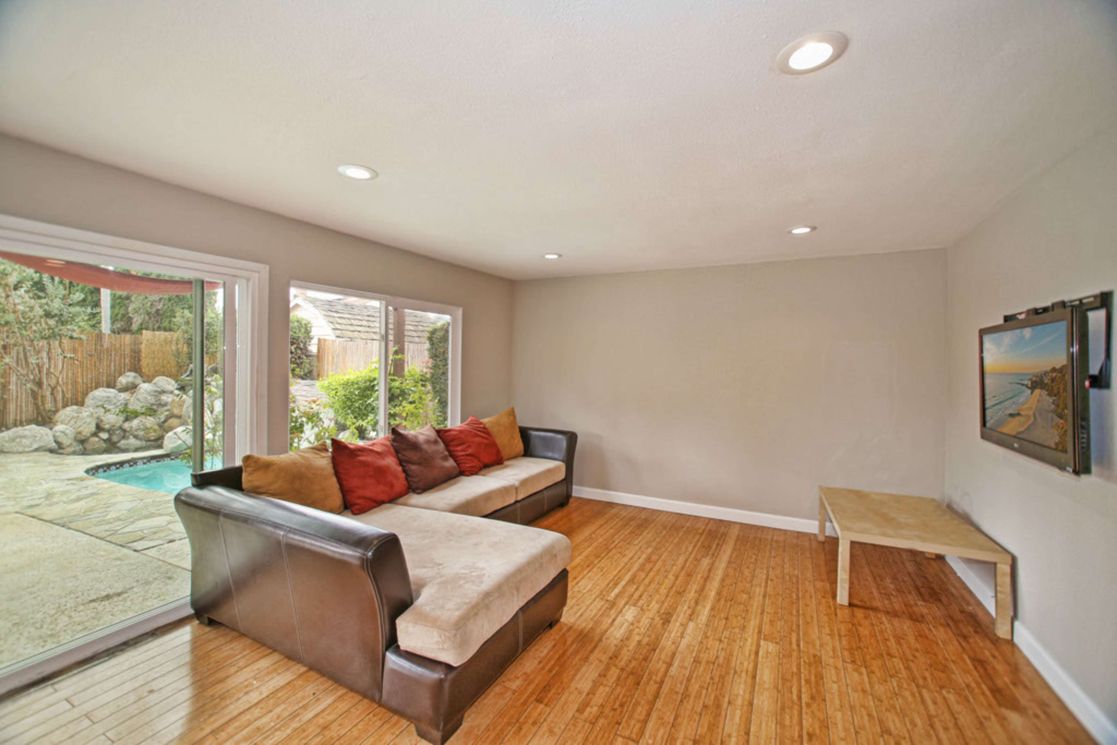 A minimalist living room features a sectional sofa with colorful pillows, a wooden coffee table, and large windows overlooking a patio and pool area.