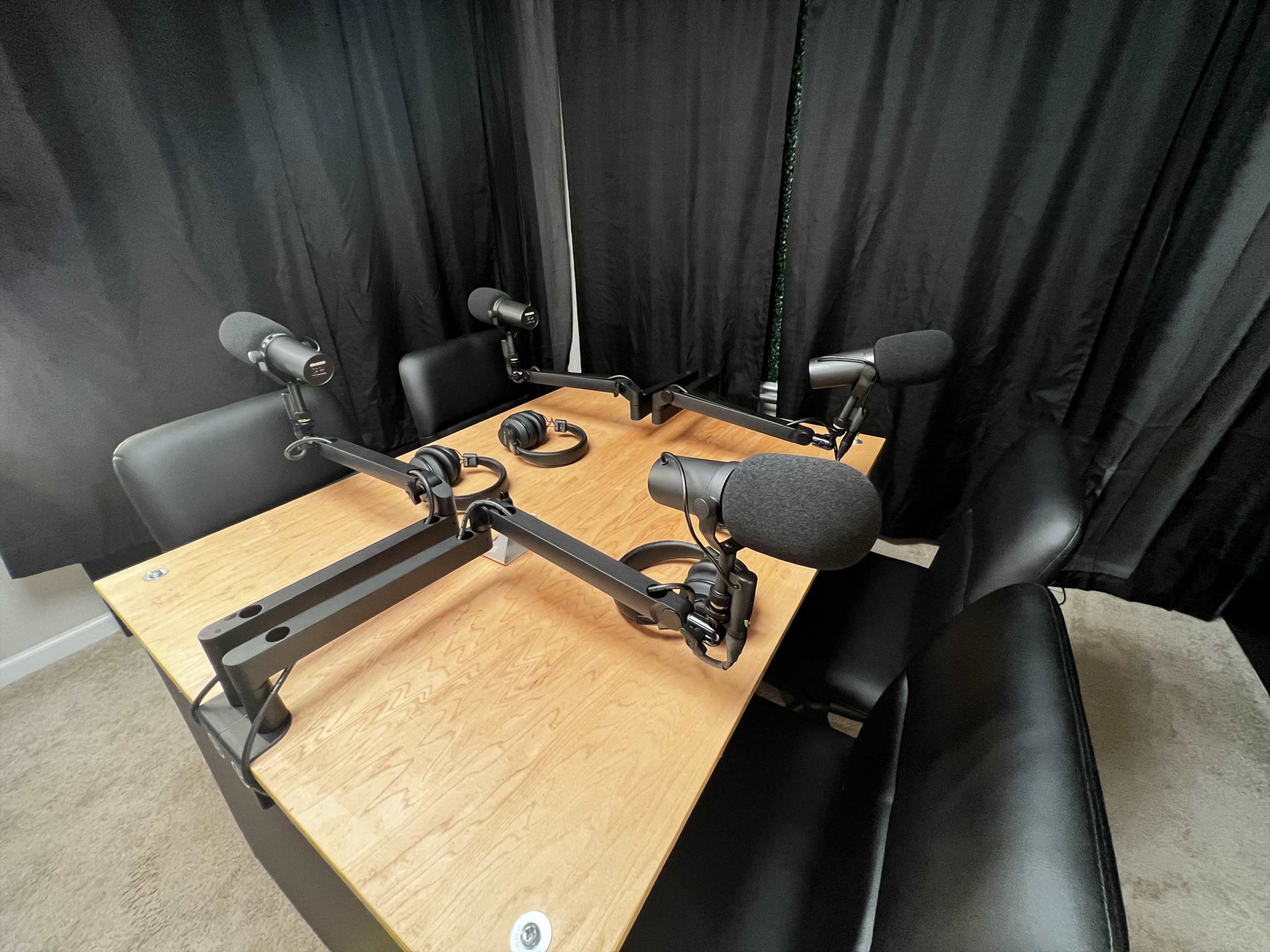 A small recording studio setup with a wooden table, four microphone stands, and black chairs, all surrounded by black curtains.