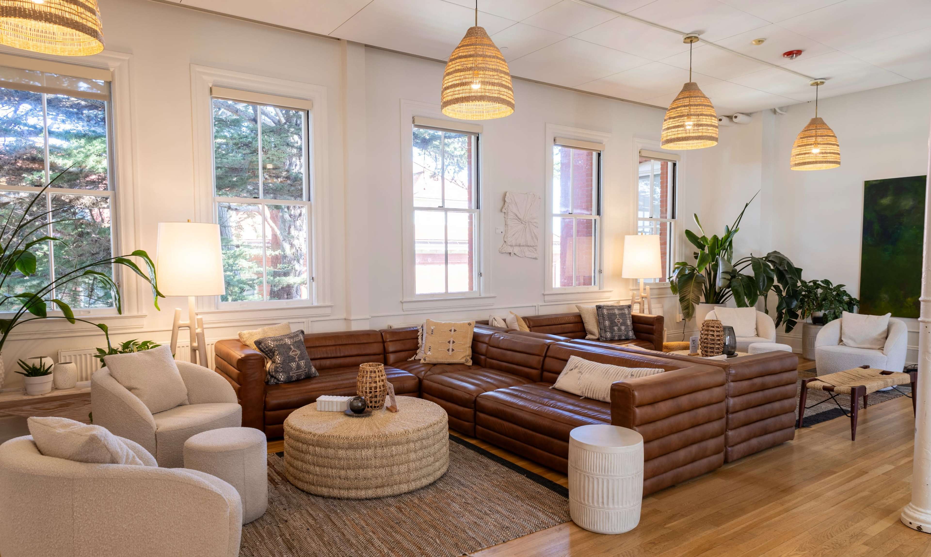 The image shows a well-lit living room featuring brown leather sofas, a round coffee table, and various plants, with large windows allowing natural light to enter.