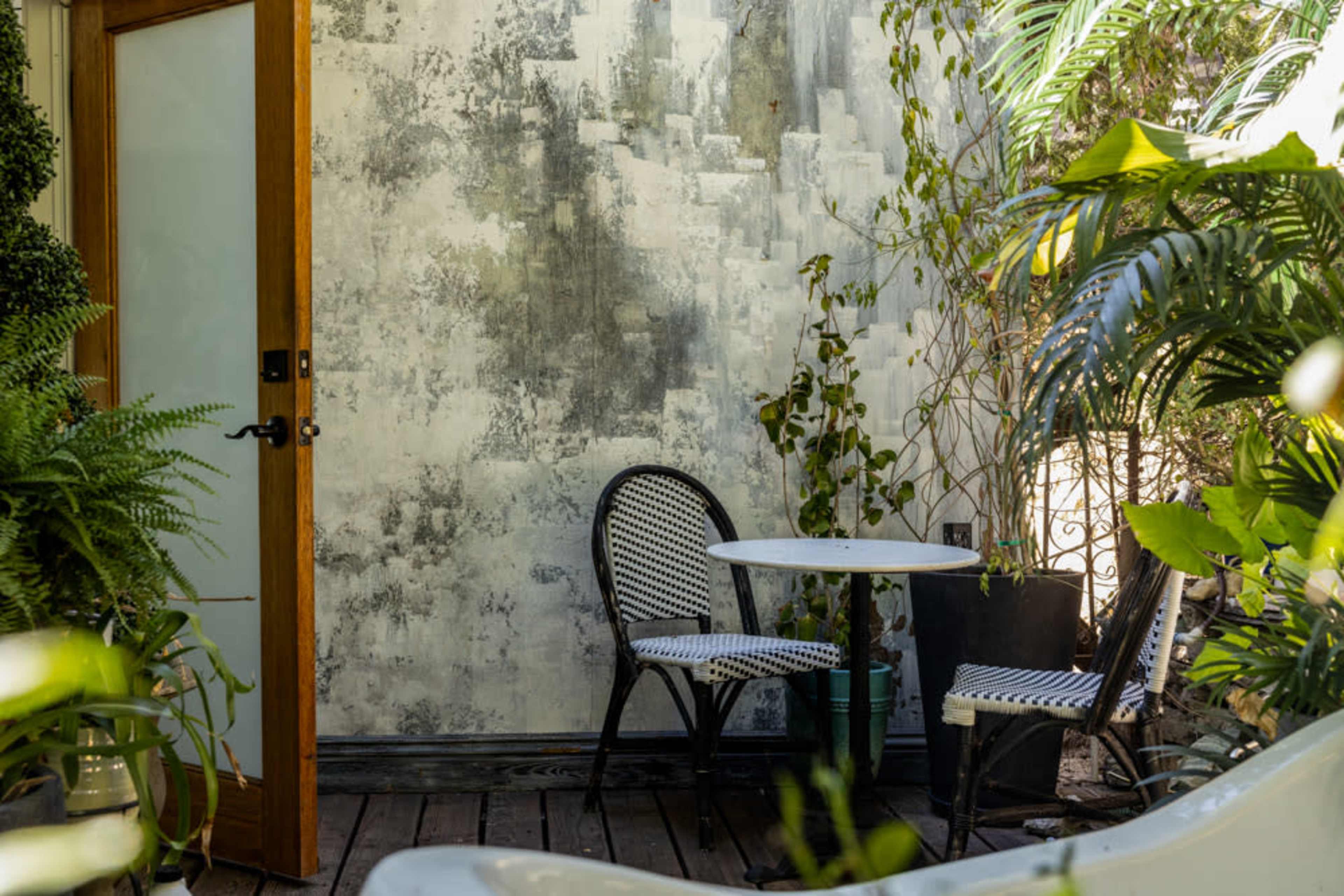 The image features a small outdoor seating area with two chairs and a round table set against a textured wall, surrounded by various plants.