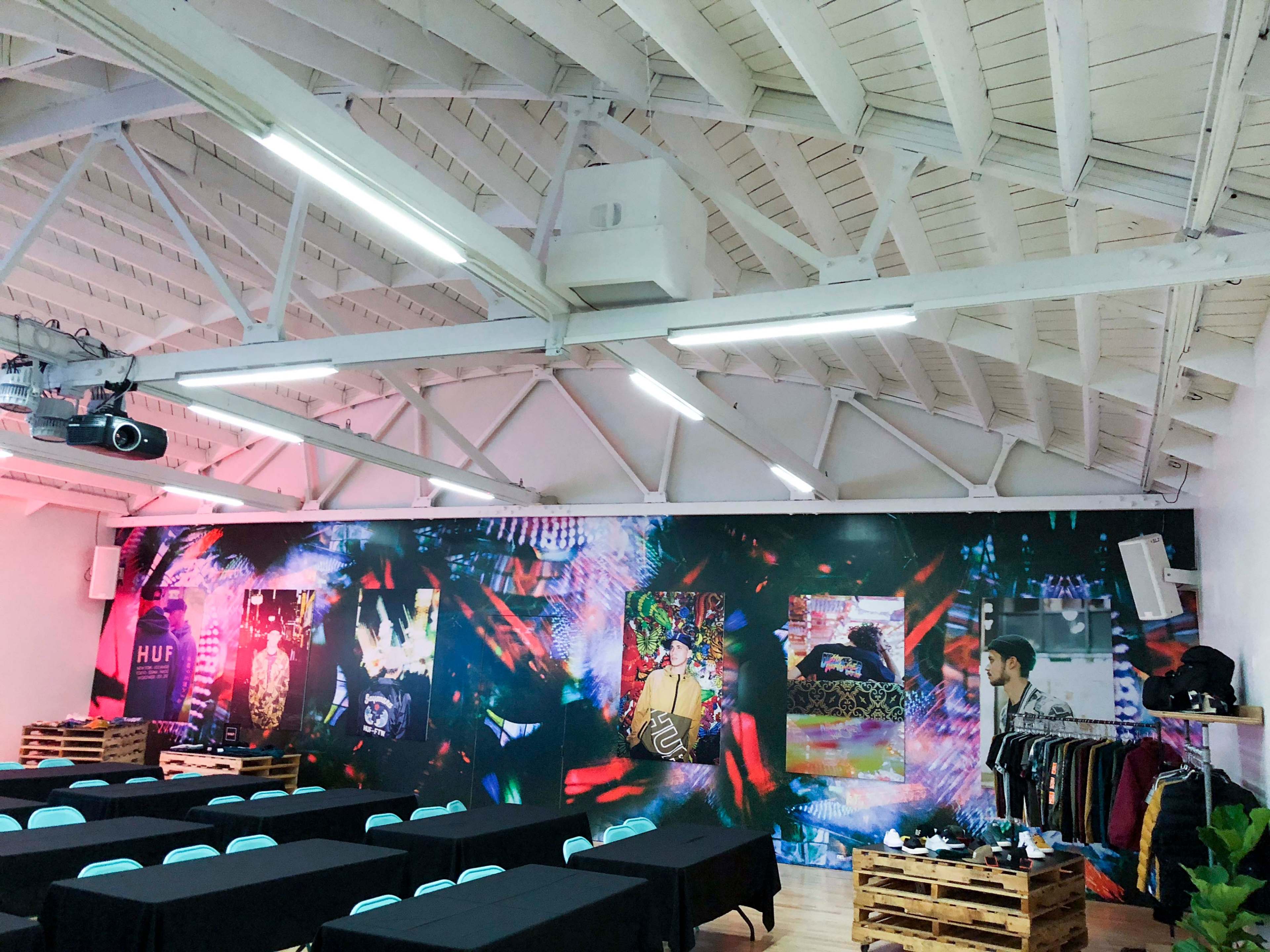 A spacious indoor venue with a colorful wall mural, rows of black tables, and a person standing near a clothing rack.