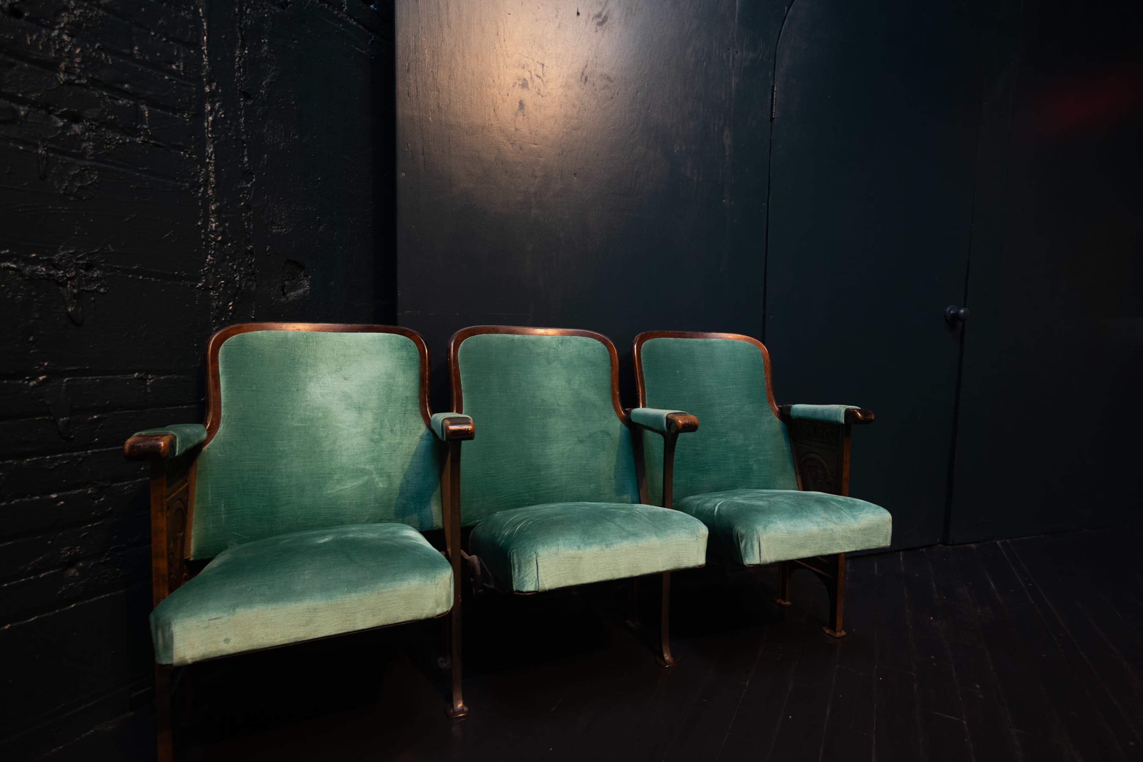 Three green upholstered chairs with wooden armrests are placed against a dark wall.