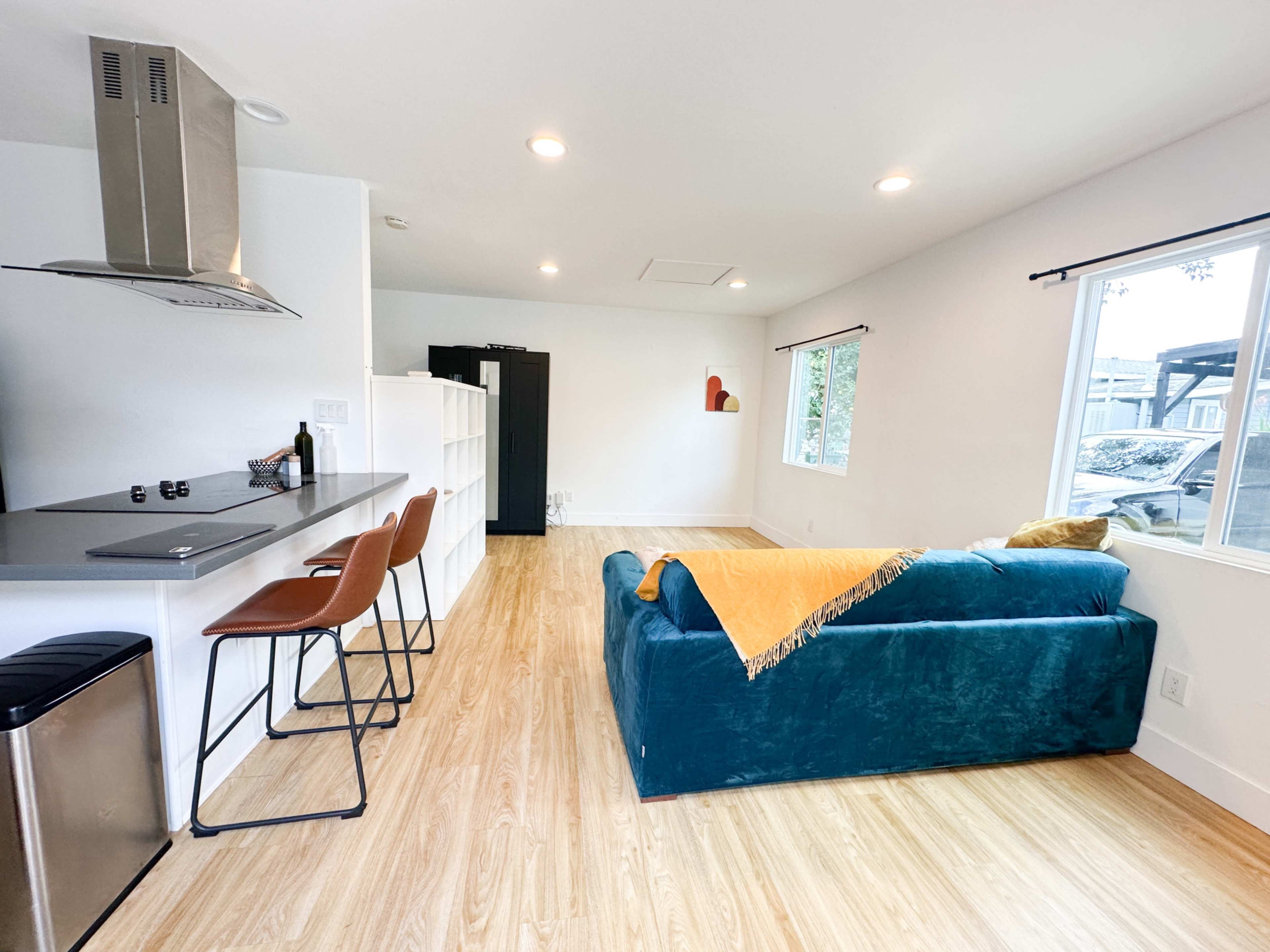 A modern living space featuring a blue sofa with an orange throw, a kitchen area with bar stools, and large windows that allow natural light.