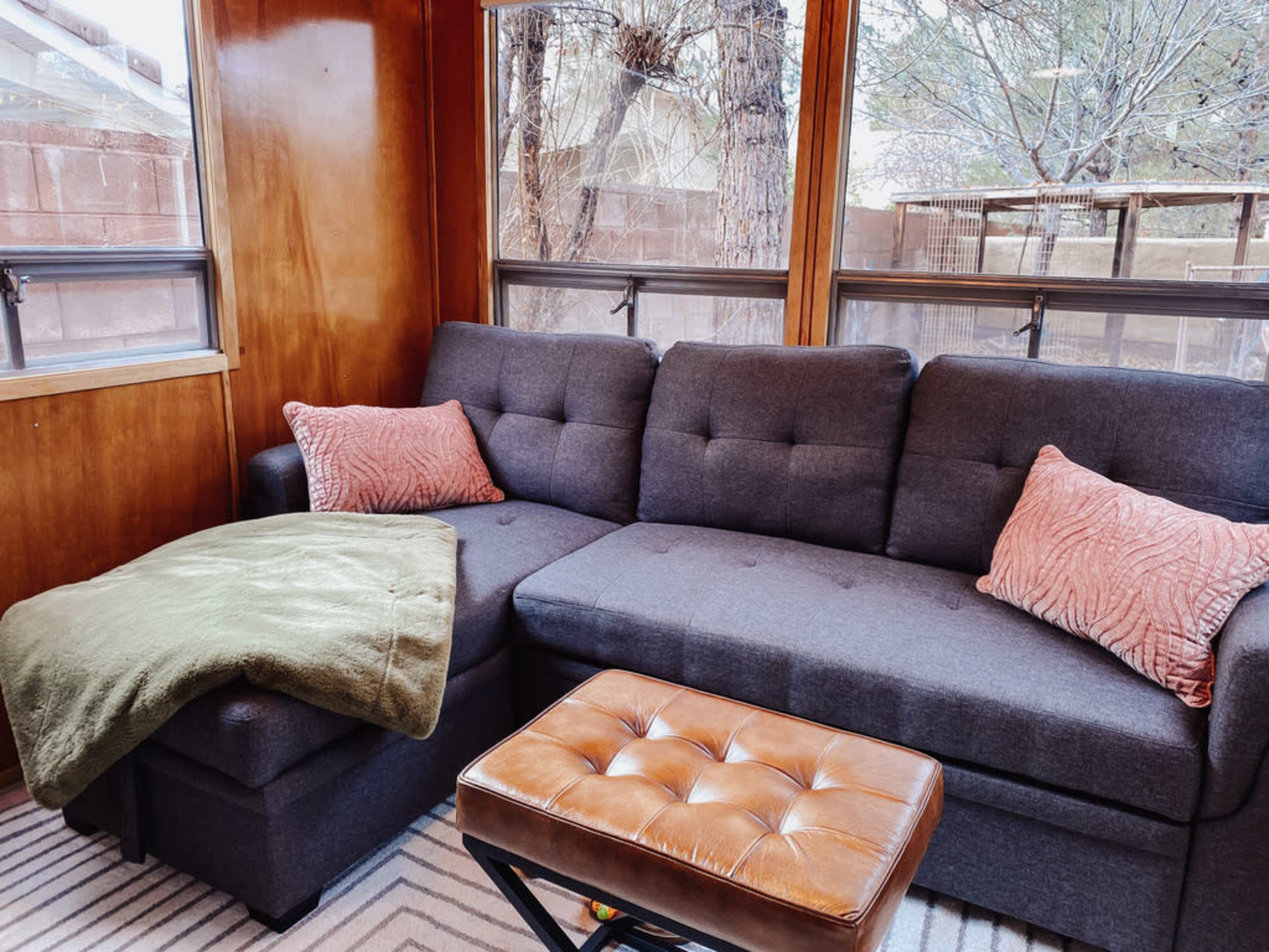 The image shows a cozy seating area with a gray sectional sofa, two cushions, a green throw blanket, and a brown tufted coffee table in a room with wooden walls and windows.