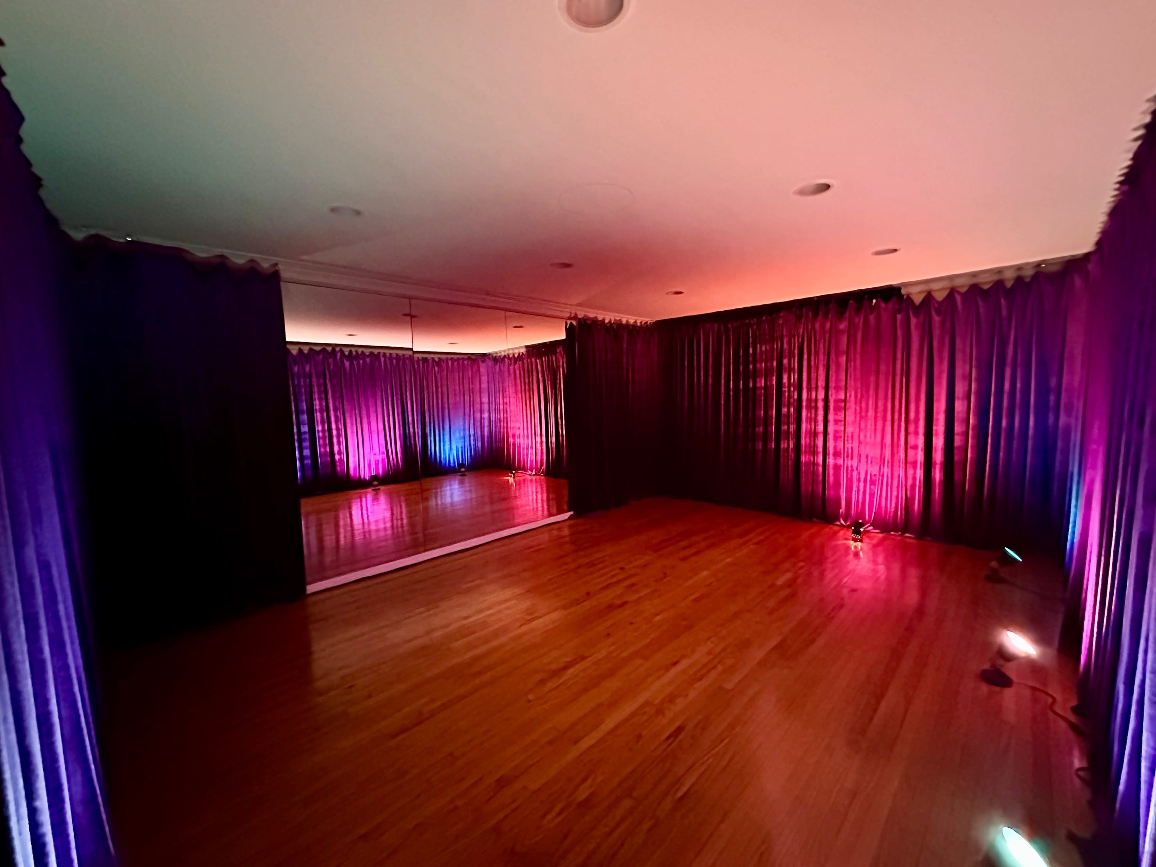 The image shows a dance studio with wooden flooring, illuminated by colored lights and bordered by purple curtains.