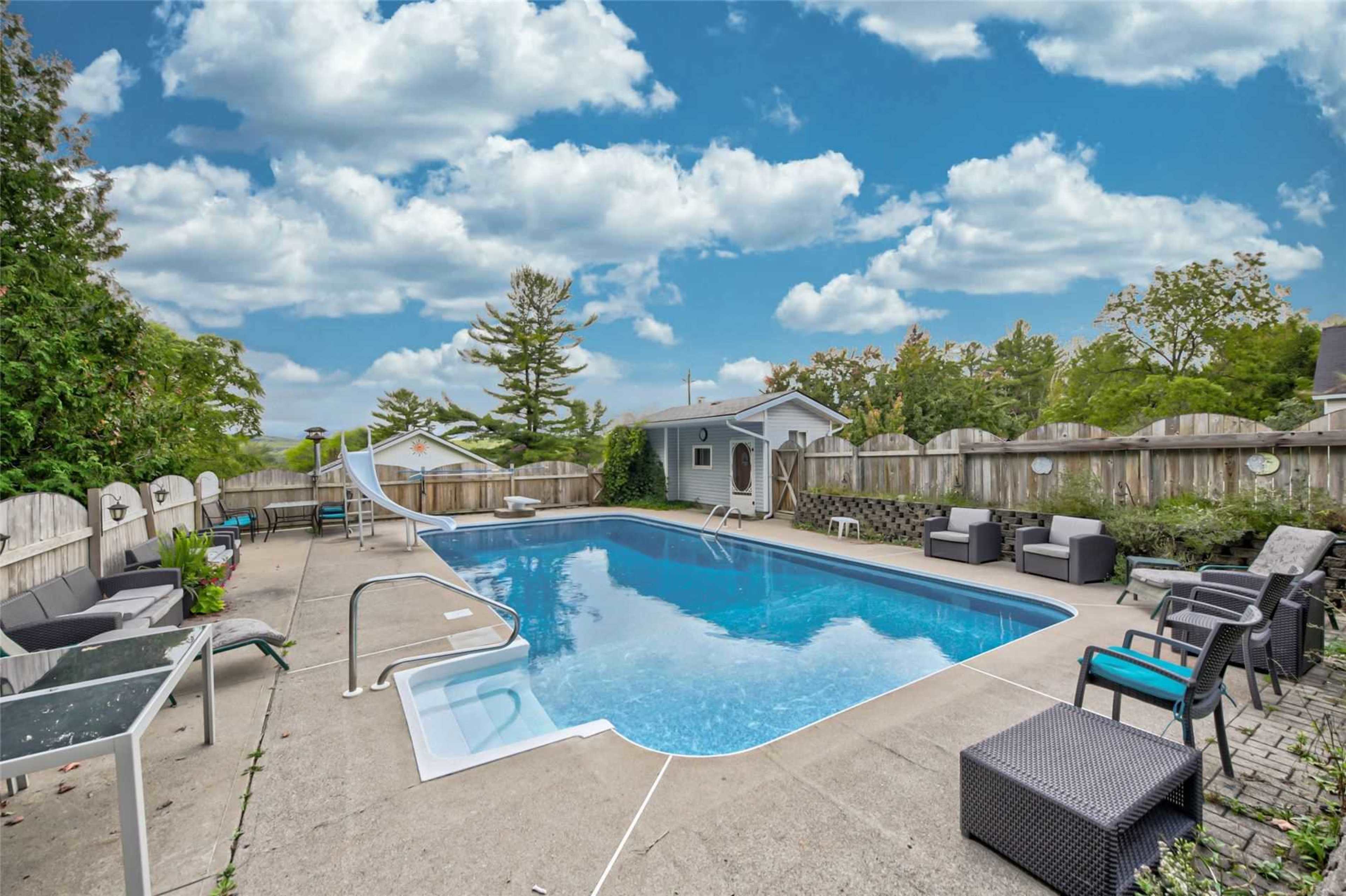 A swimming pool surrounded by lounge chairs and a slide, with a fence and trees in the background.
