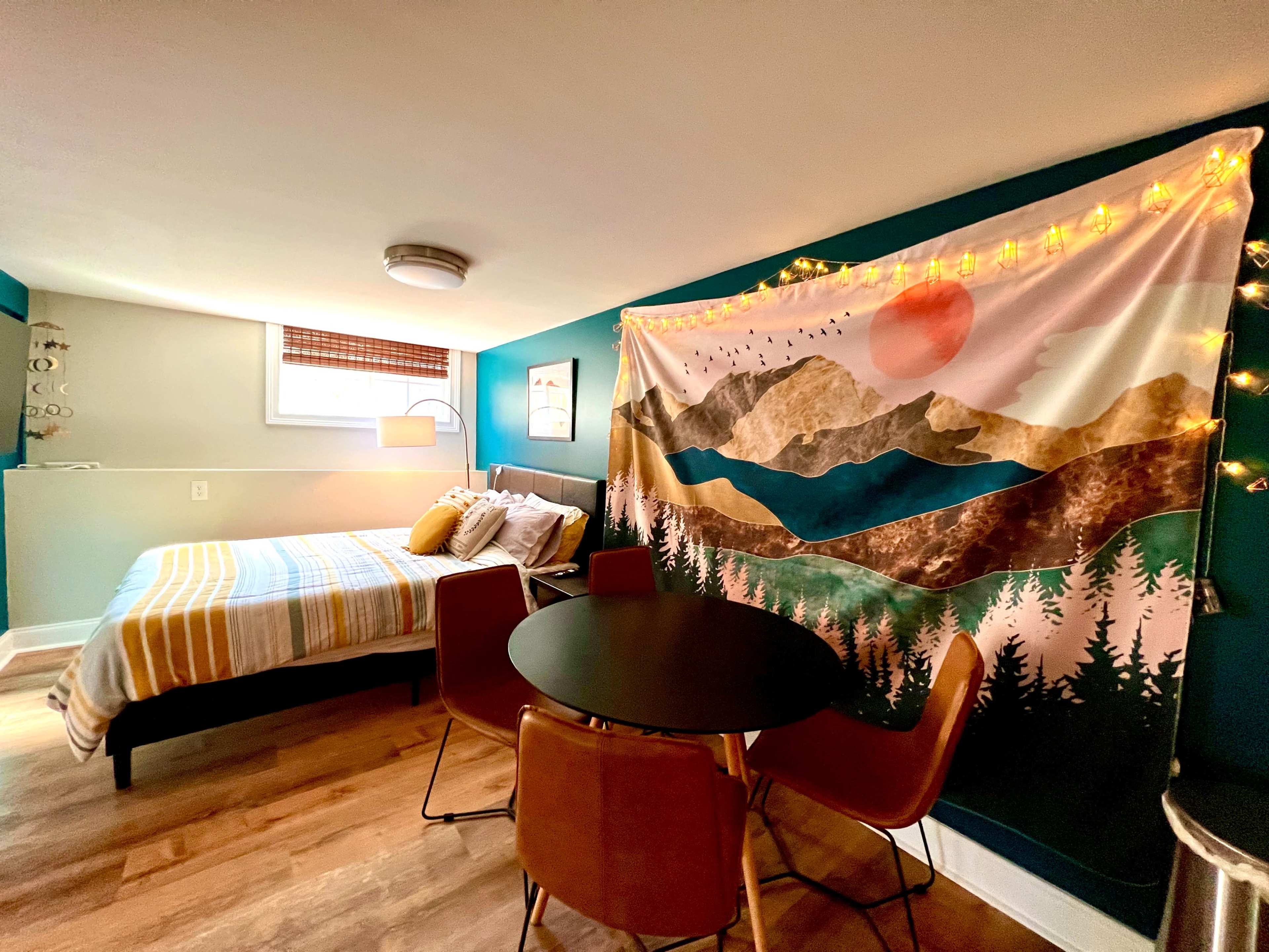 A cozy room features a bed with striped bedding, a round dining table with four chairs, and a tapestry depicting mountains and a sunset hanging on the wall.