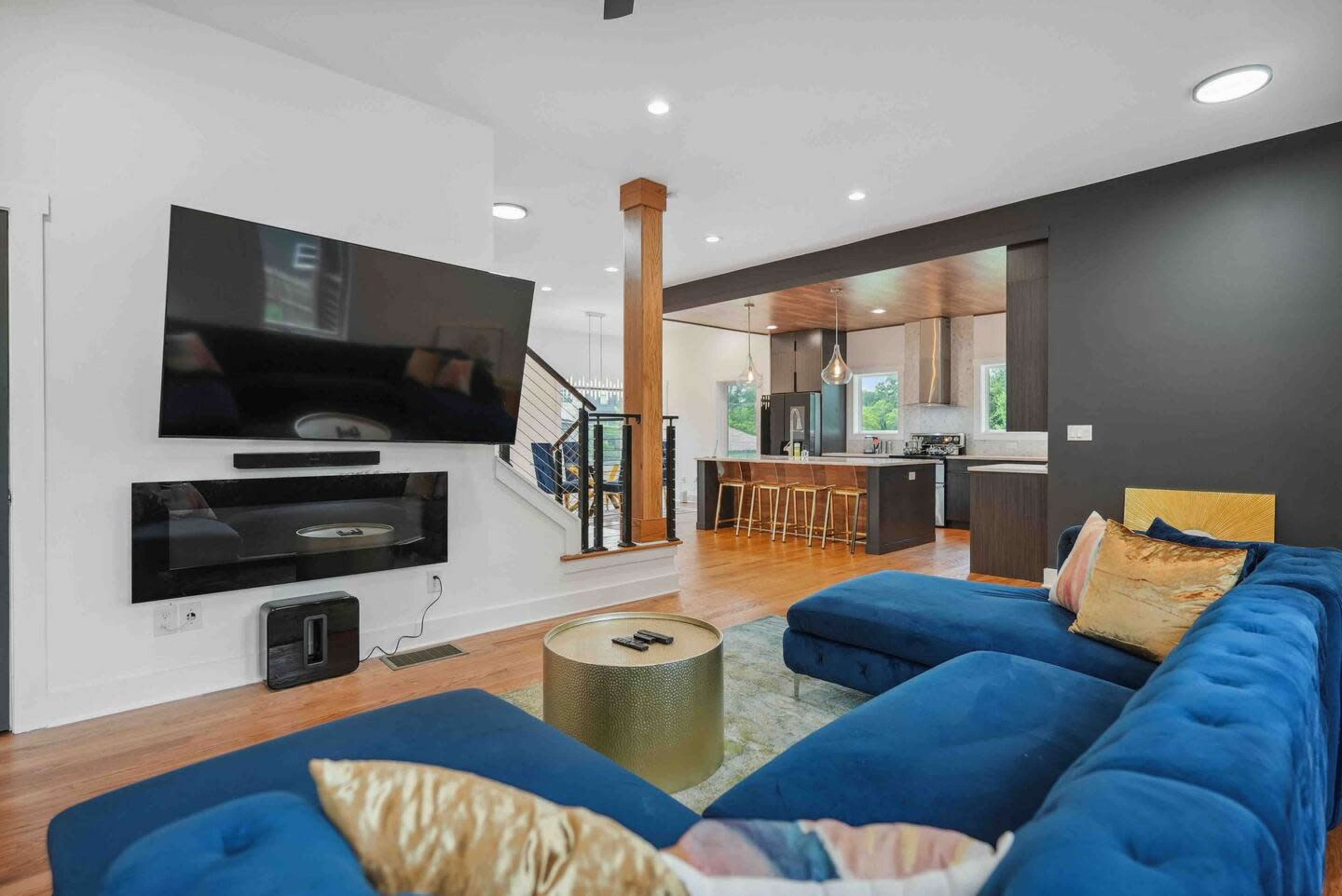 A modern living room featuring a blue sectional sofa, a large flat-screen TV mounted on the wall, and a kitchen area visible in the background with wooden bar stools.