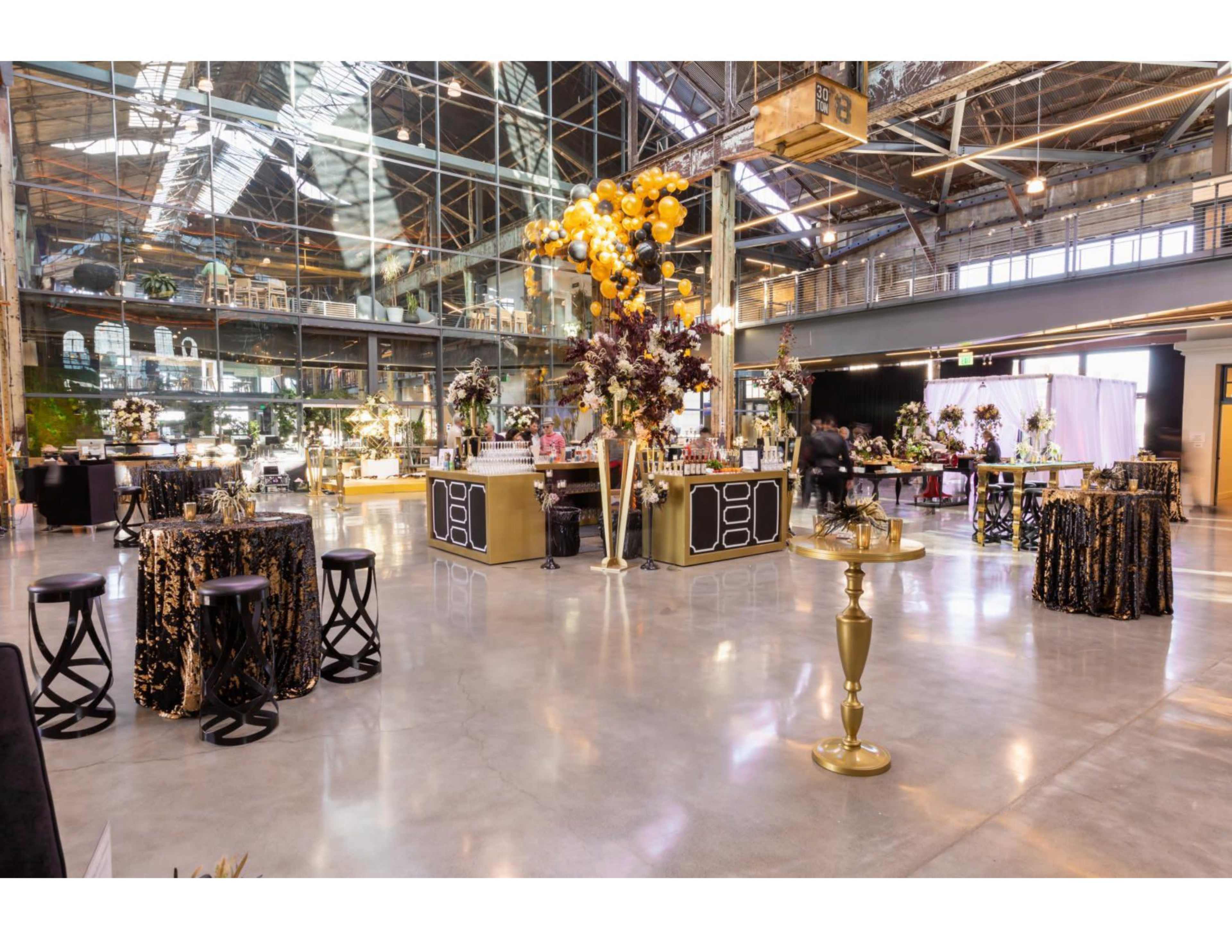 An elegantly decorated interior space for an event, featuring black and gold furnishings, floral arrangements, and a bar.
