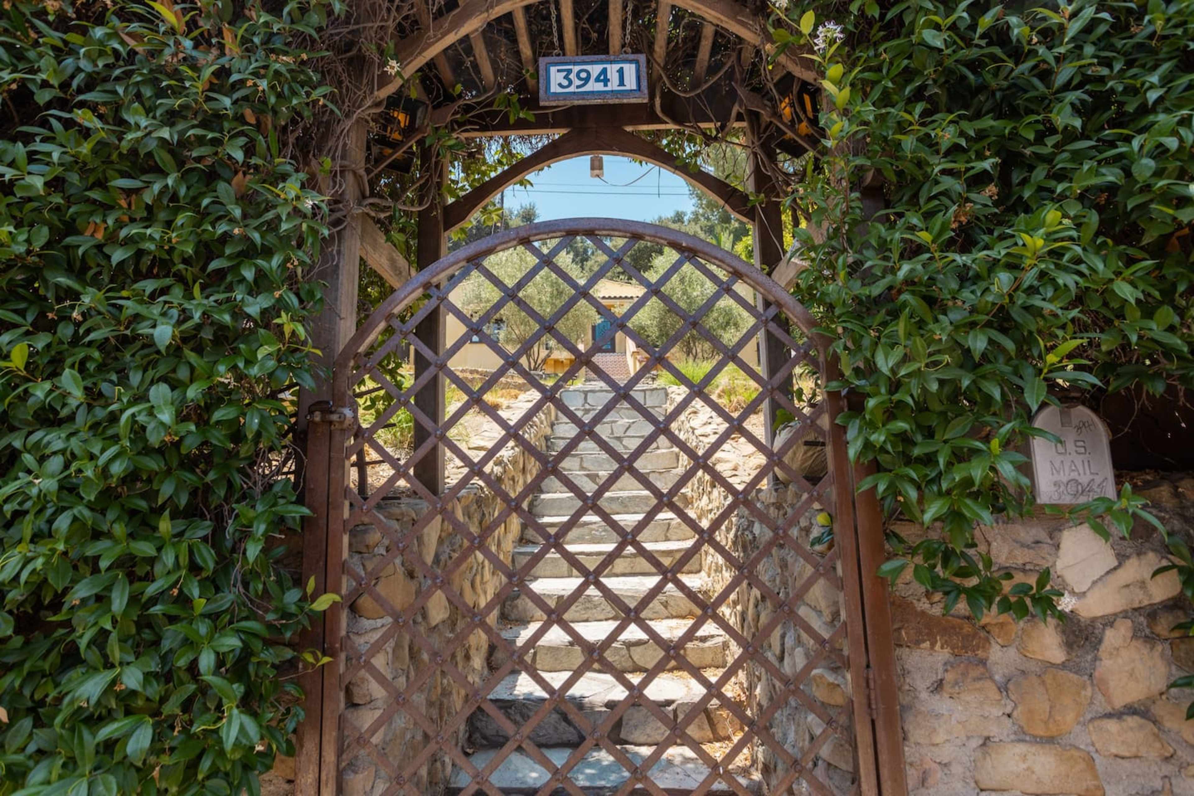A wrought iron gate with a diamond pattern leads into a stone pathway bordered by greenery, with a house visible in the background.