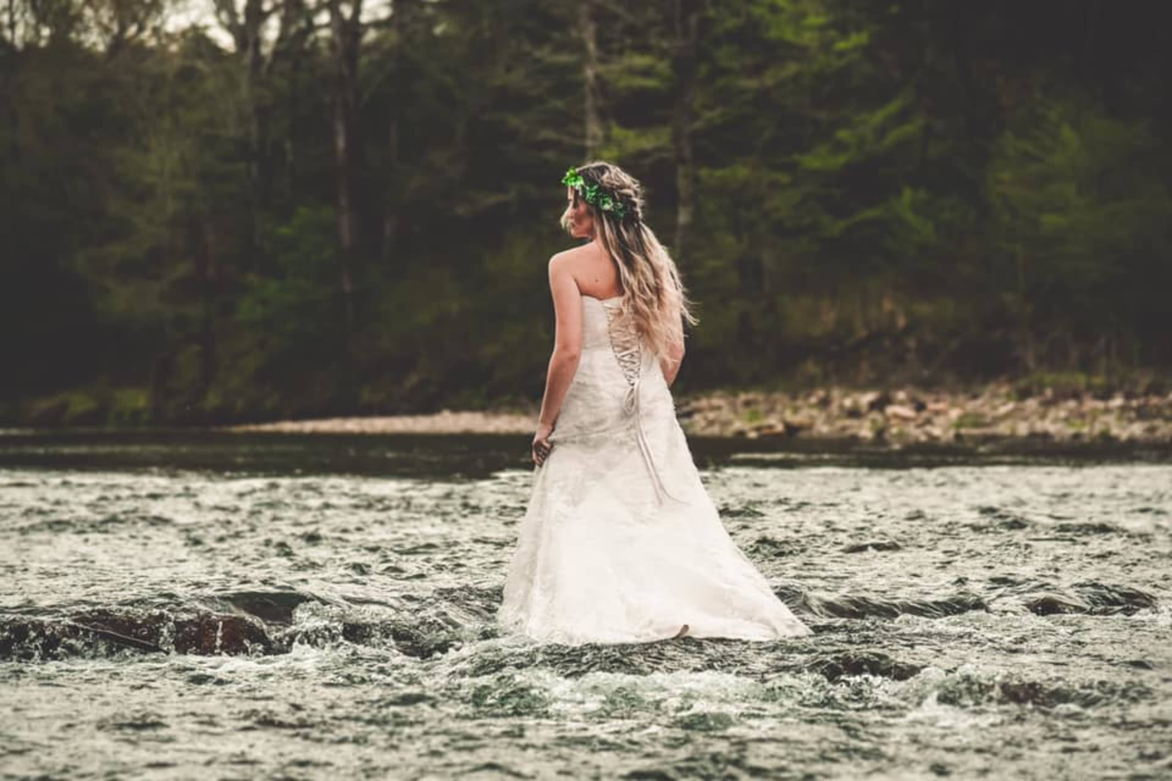 A woman in a wedding dress stands barefoot in a shallow river, surrounded by trees.