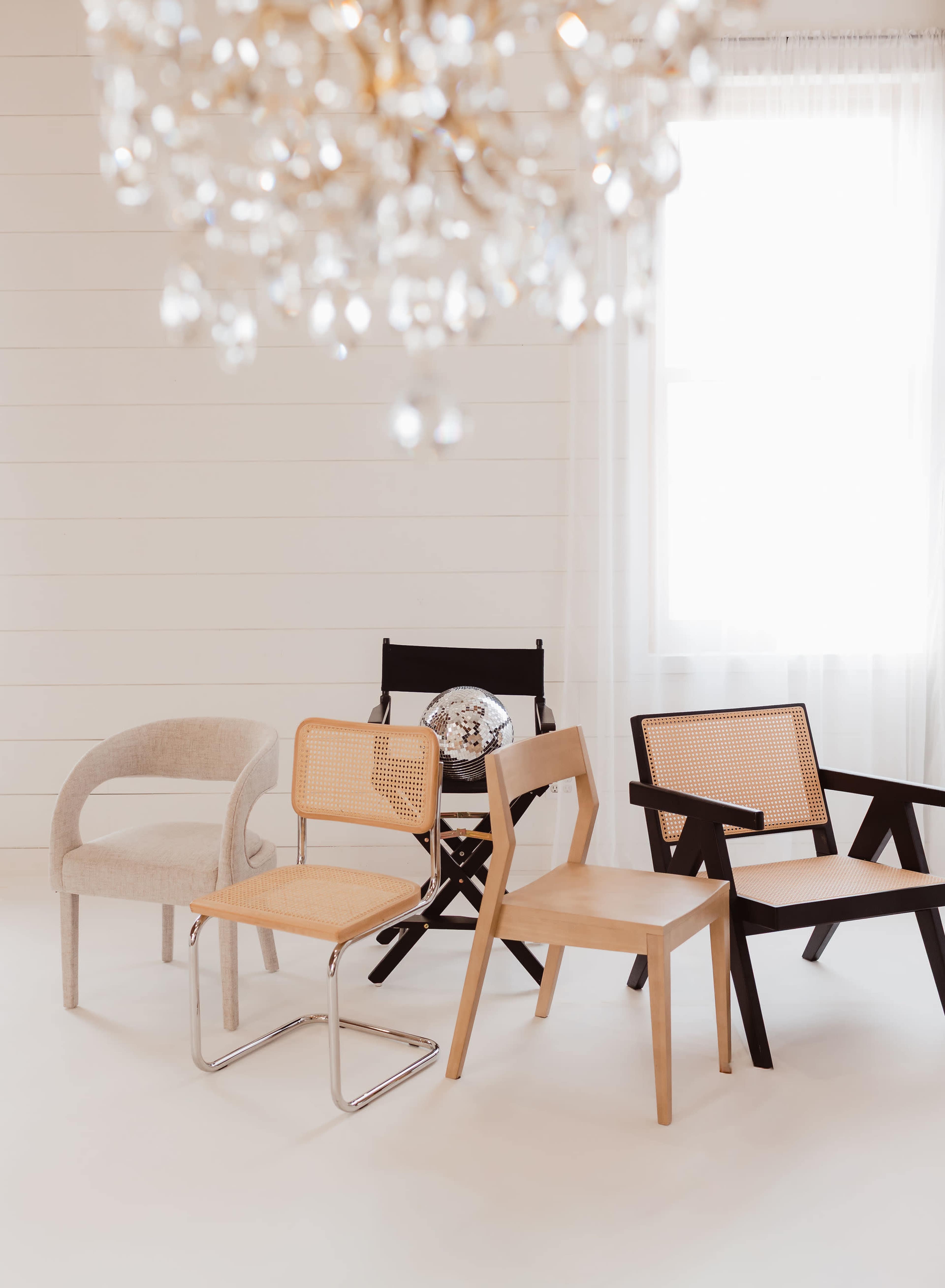 The image features four different styles of chairs arranged in a well-lit, minimalistic room with a chandelier overhead.