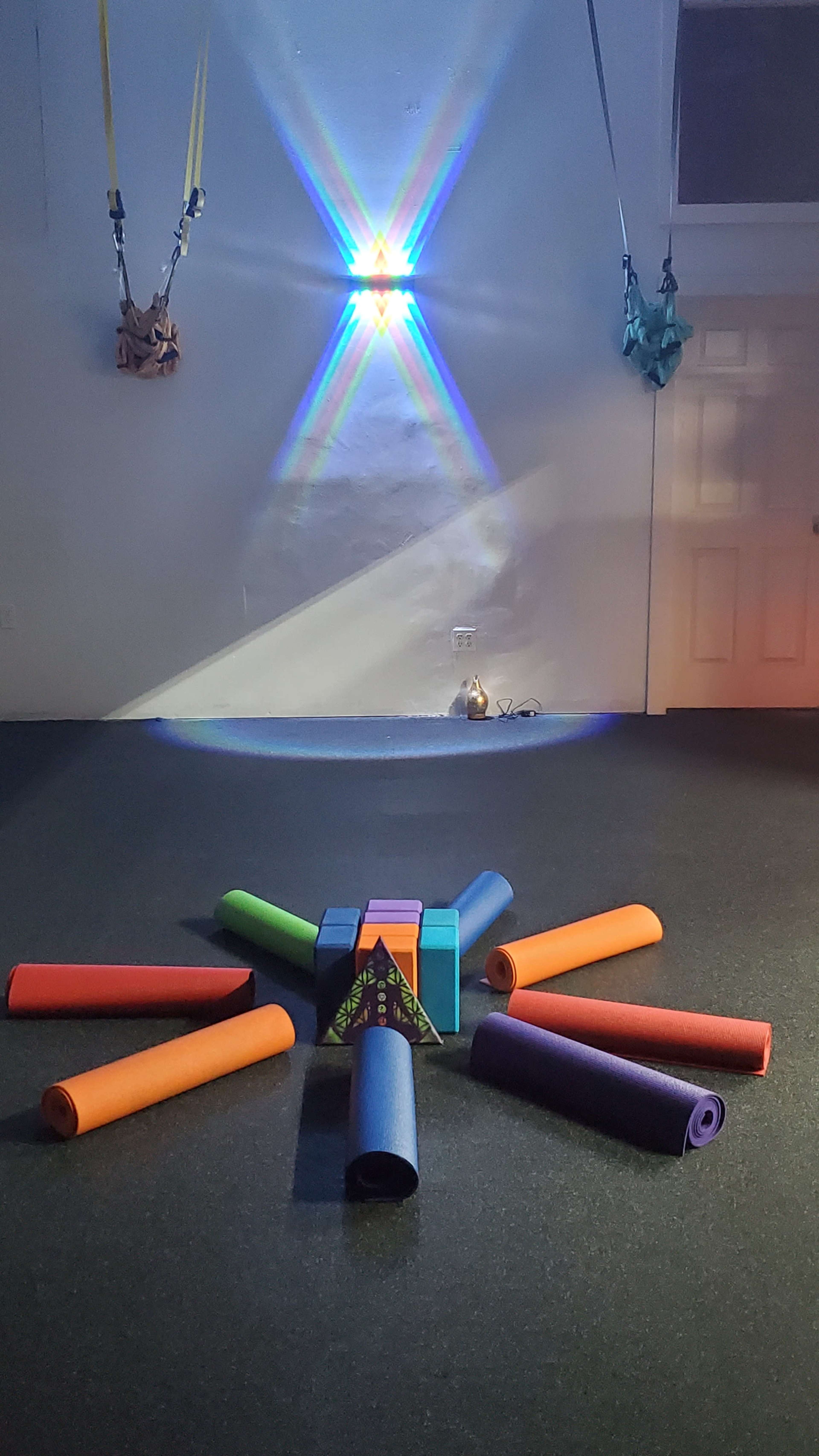 A fitness studio with colorful exercise mats arranged in a circular pattern around a small pyramid-shaped structure, illuminated by a light projection on the wall.