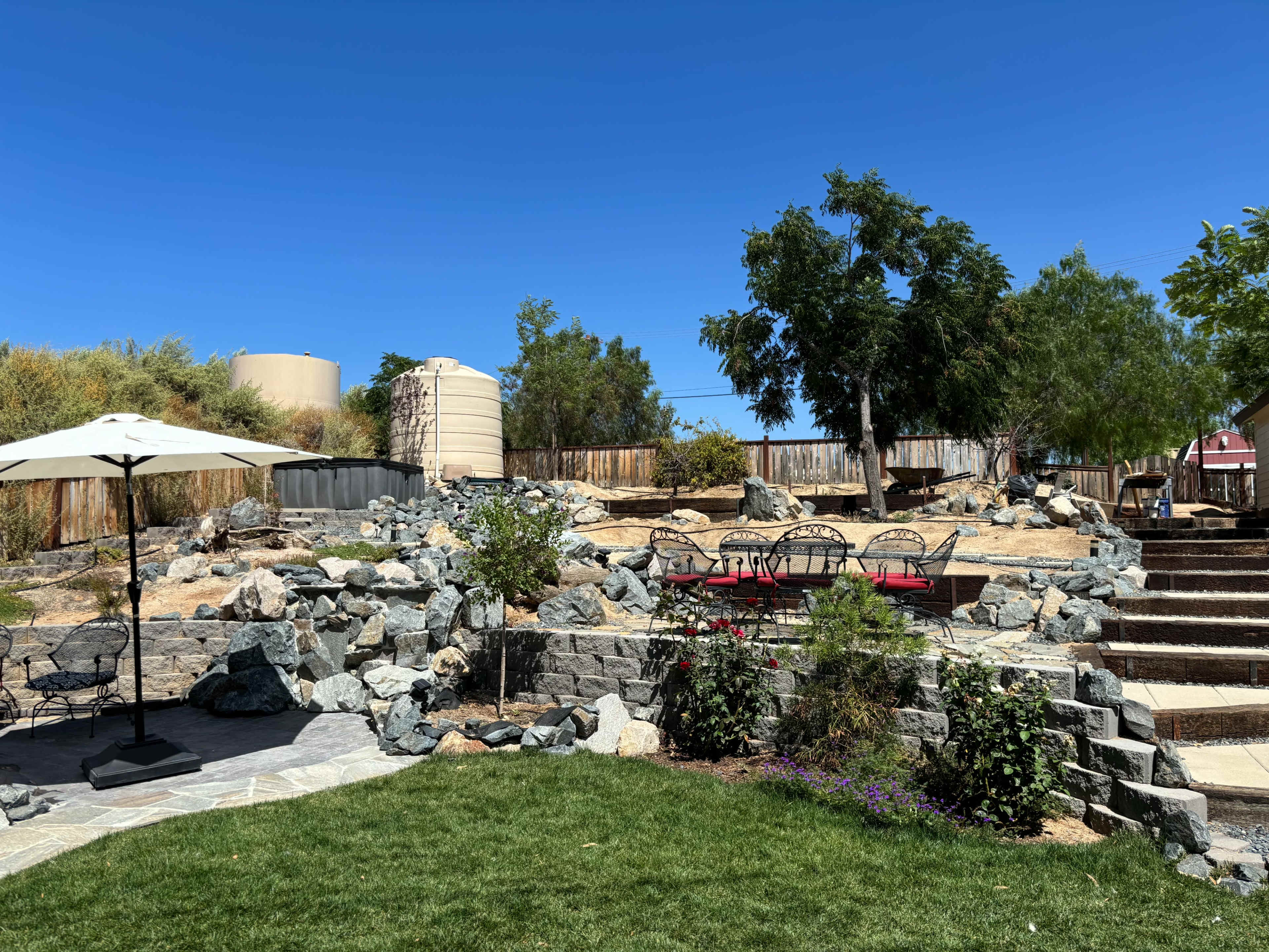 A landscape with a stone pathway leading to a rocky area, surrounded by greenery and a few trees, with an umbrella-covered seating area in the foreground.