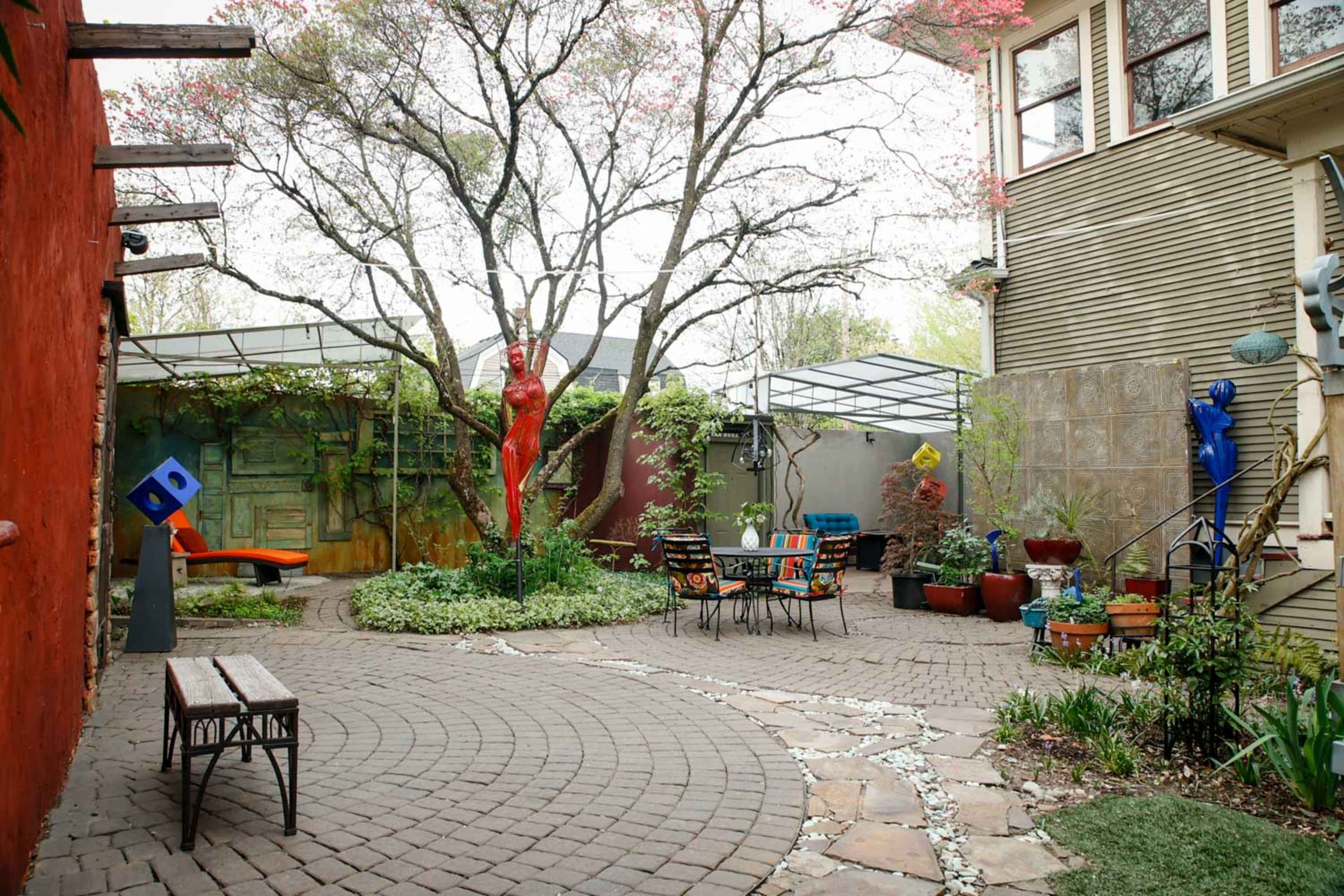 A paved courtyard features a tree, colorful sculptures, and seating areas surrounded by plants and walls of different textures.
