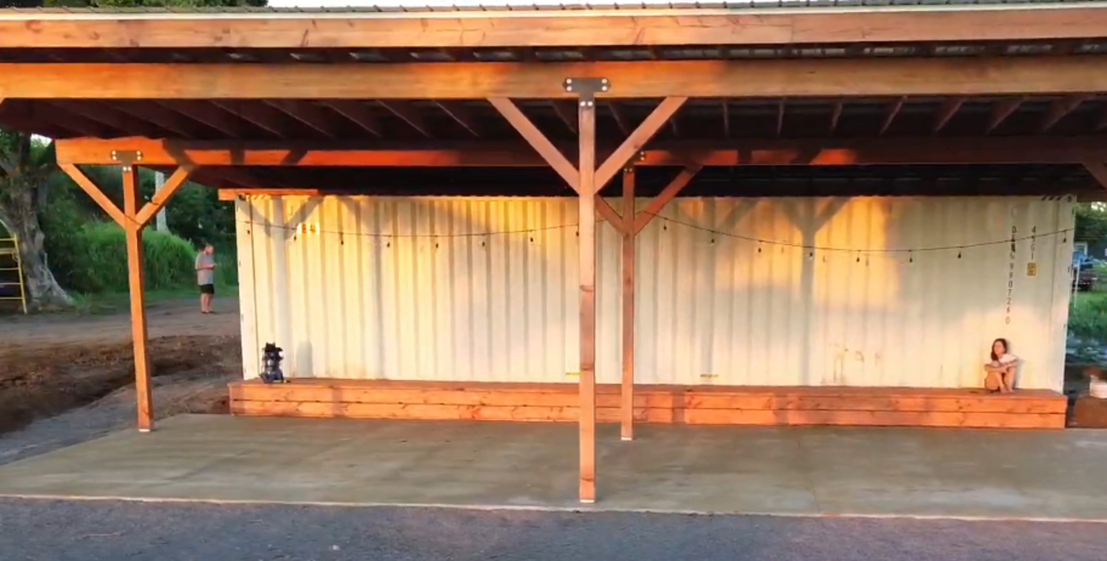 A wooden structure with a roof shelters a shipping container, while a child sits on the concrete floor beside it.