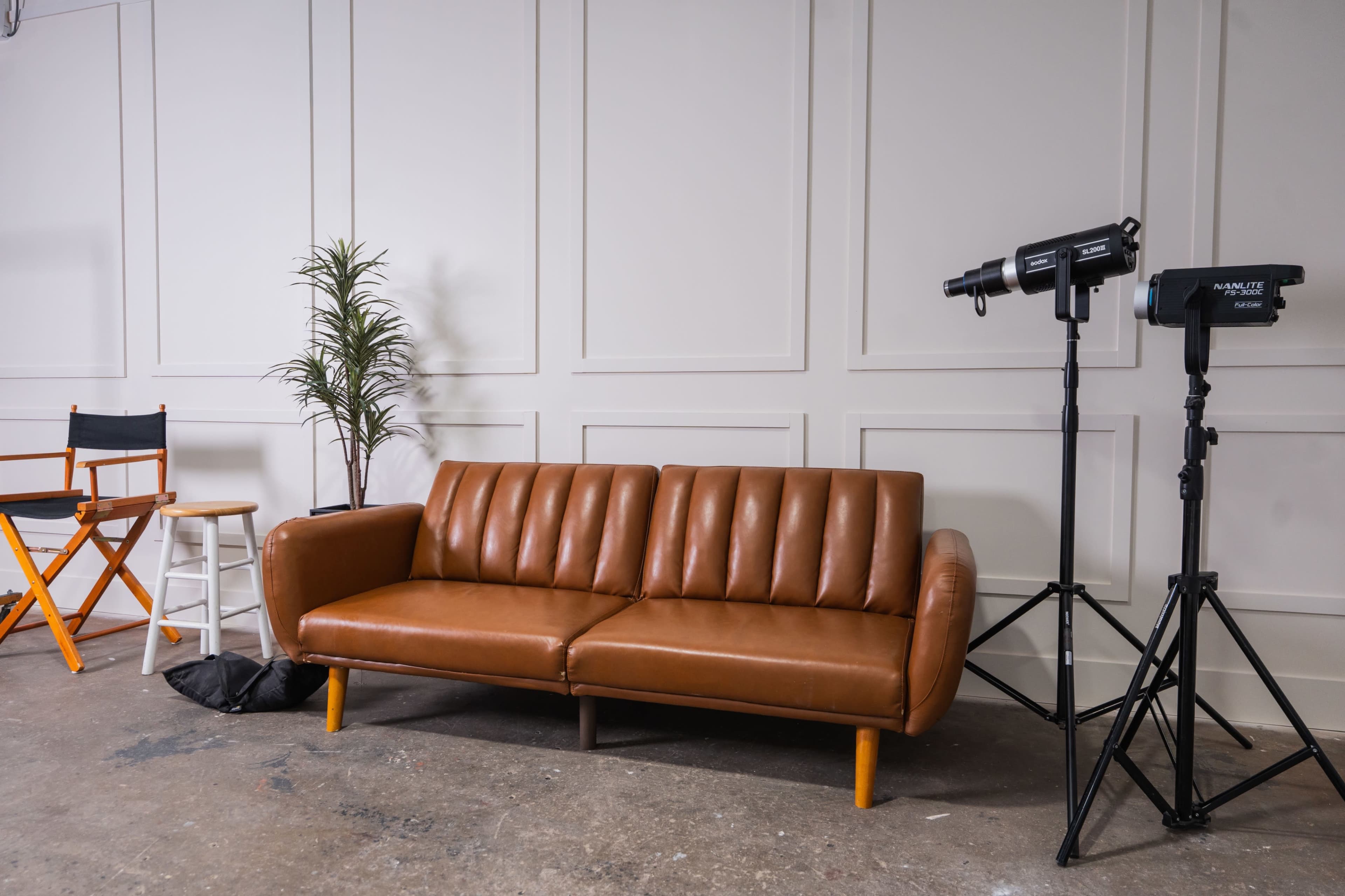 The image shows a cozy brown leather sofa positioned against a white wall, accompanied by a potted plant, a director's chair, and two studio lights on tripods.