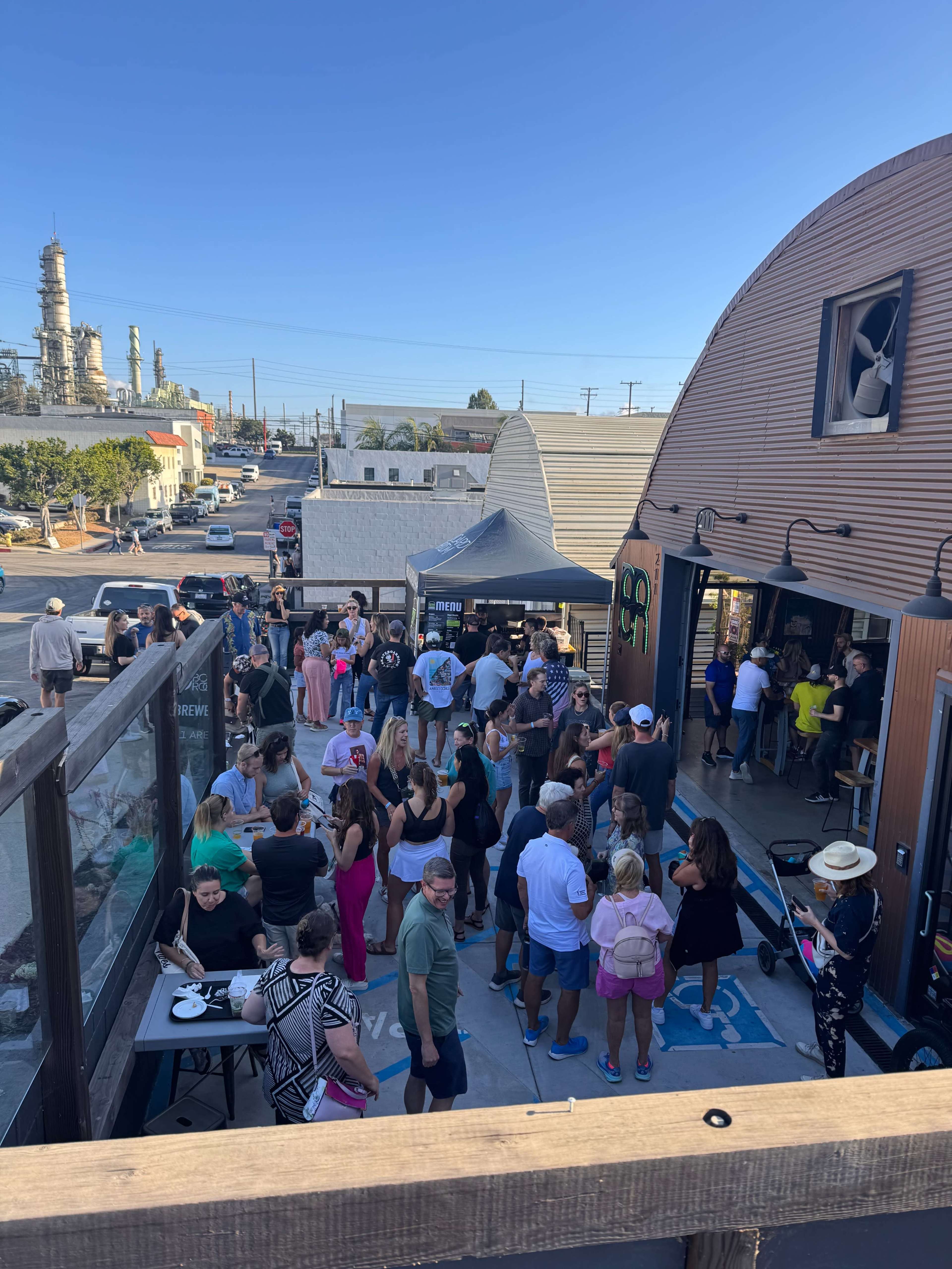A crowd of people gathers outside a bustling café with industrial buildings in the background.