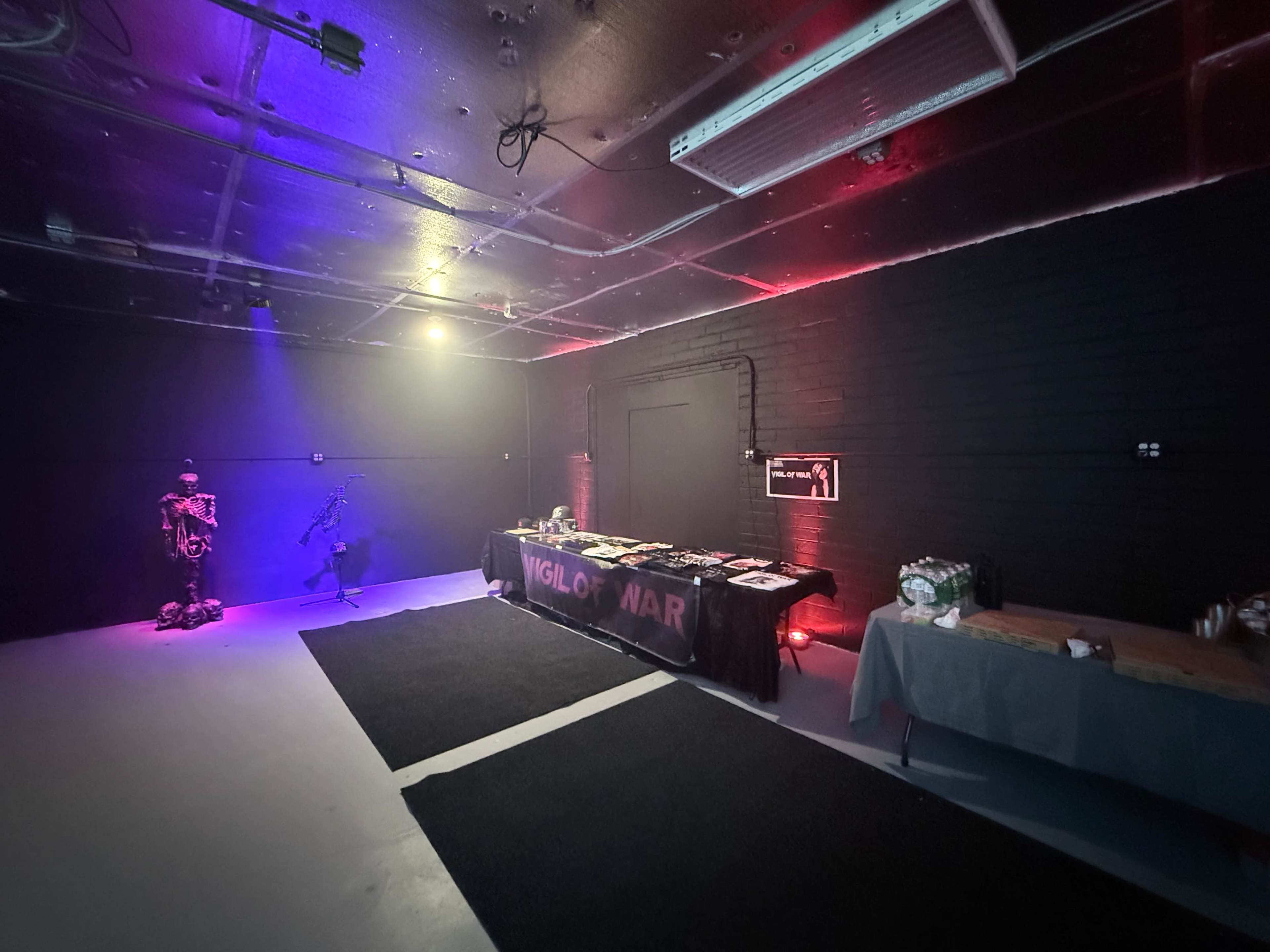 The image shows a dimly lit room with dark walls, featuring a table with various items set up in the center and a decorative skeleton figure to the left.